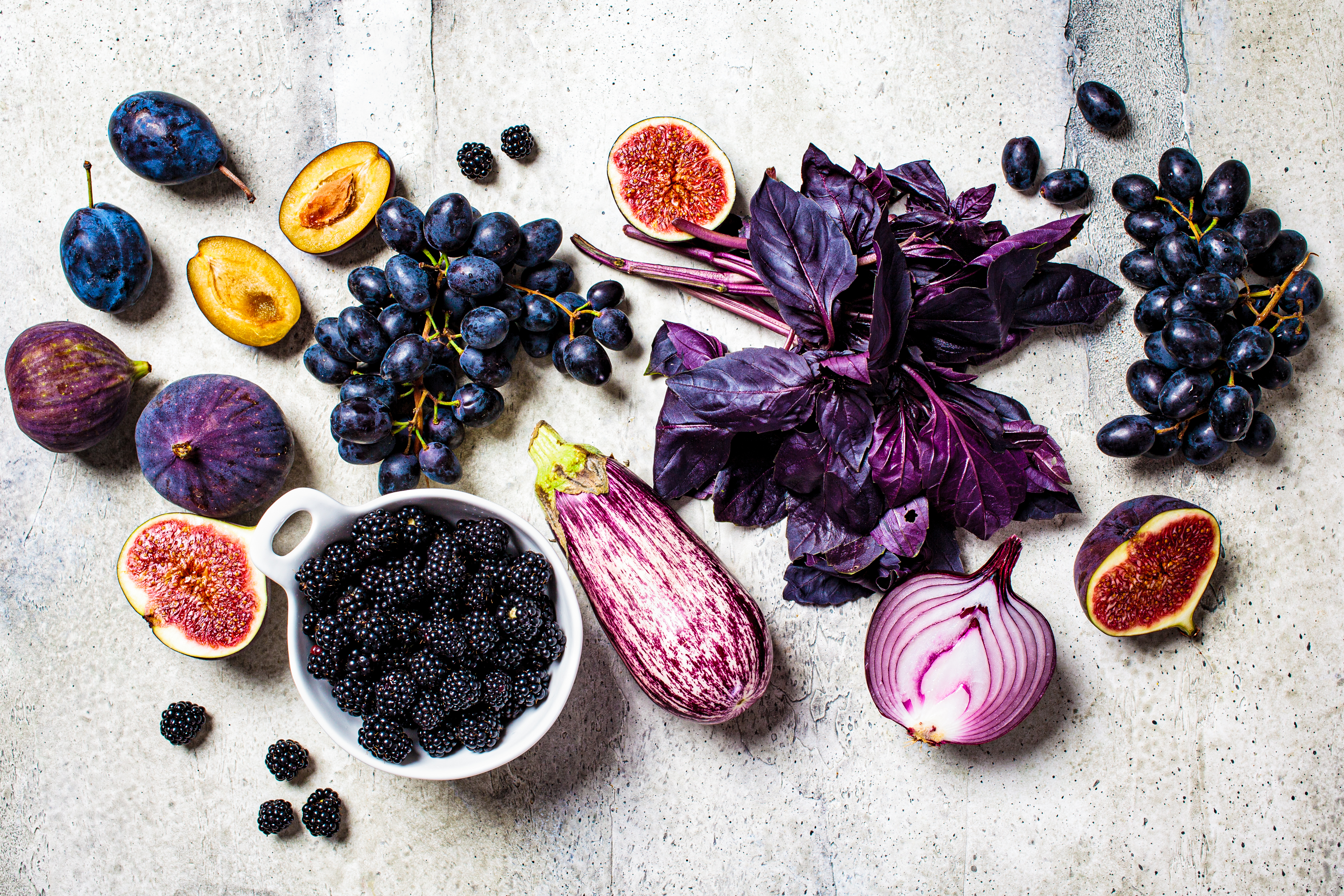 Raw purple vegetables and fruits on gray concrete background. Flat lay of purple food. Eggplant, grapes, figs, plums, blackberries, onions and basil, top view.