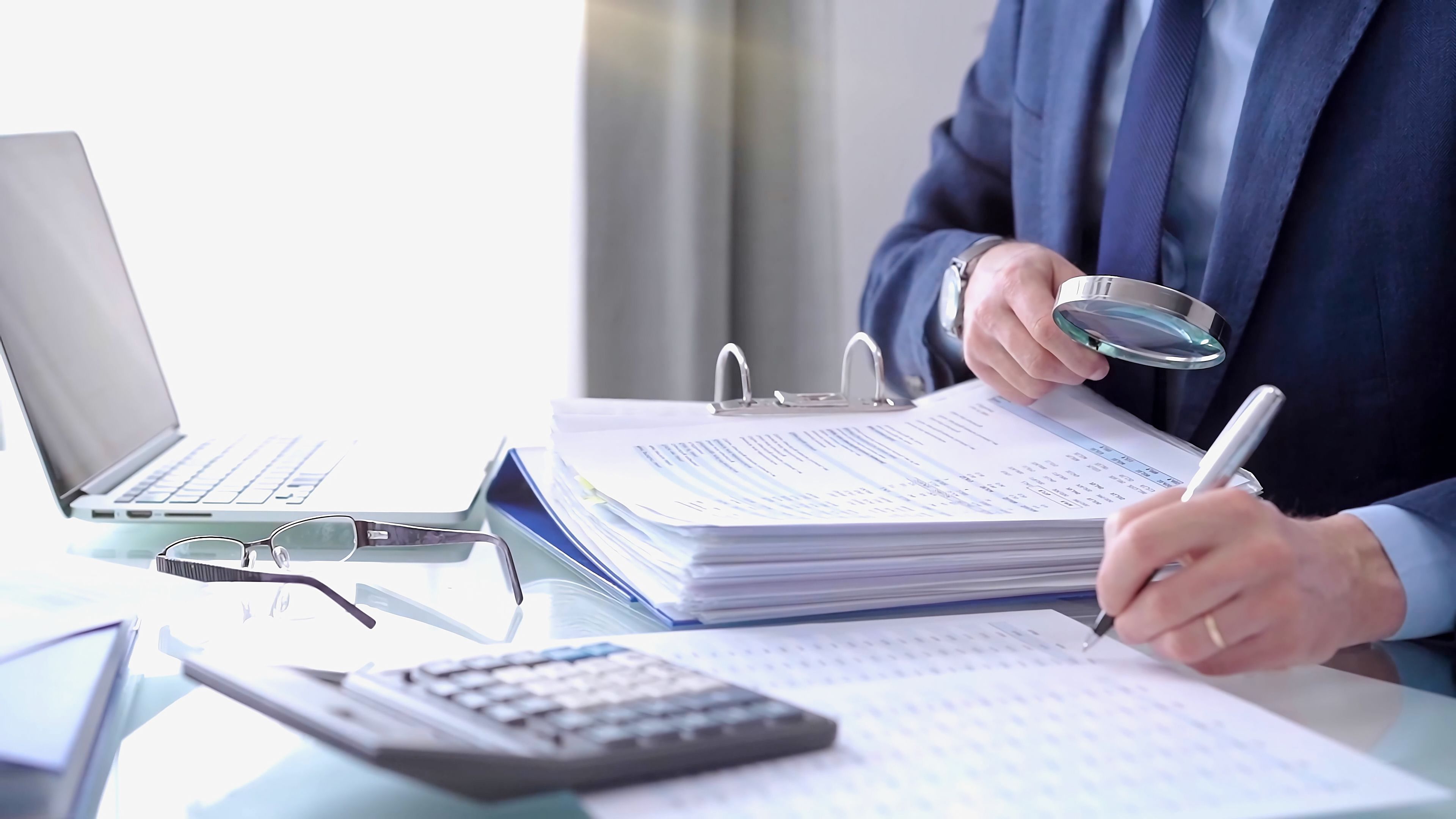 Businessman using a calculator and magnifying glass while analyzing financial documents at desk in fair modern office. Audit and taxes in business