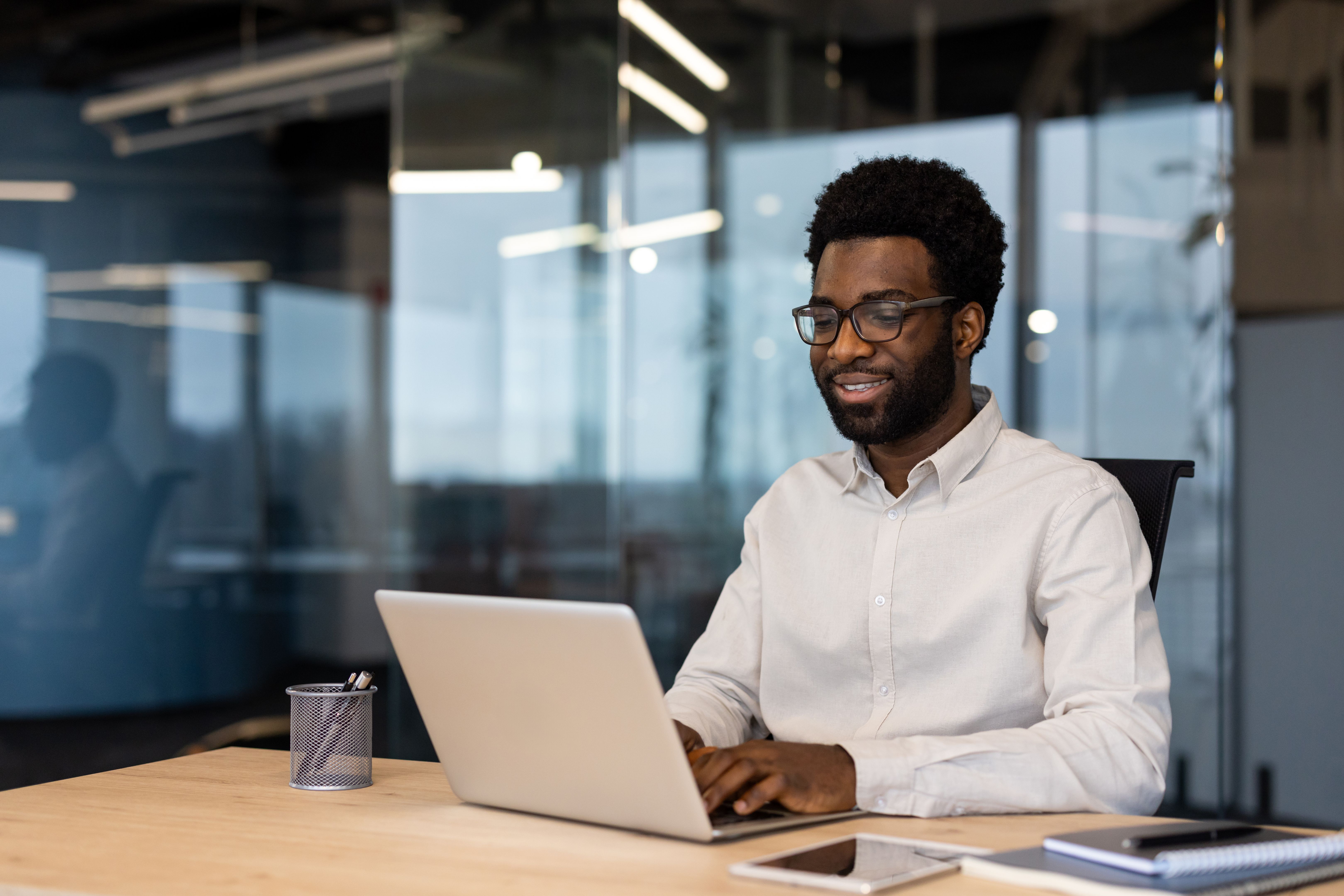 Young successful businessman working on laptop inside office, man in light shirt smiling contentedly, typing on laptop keyboard at workplace