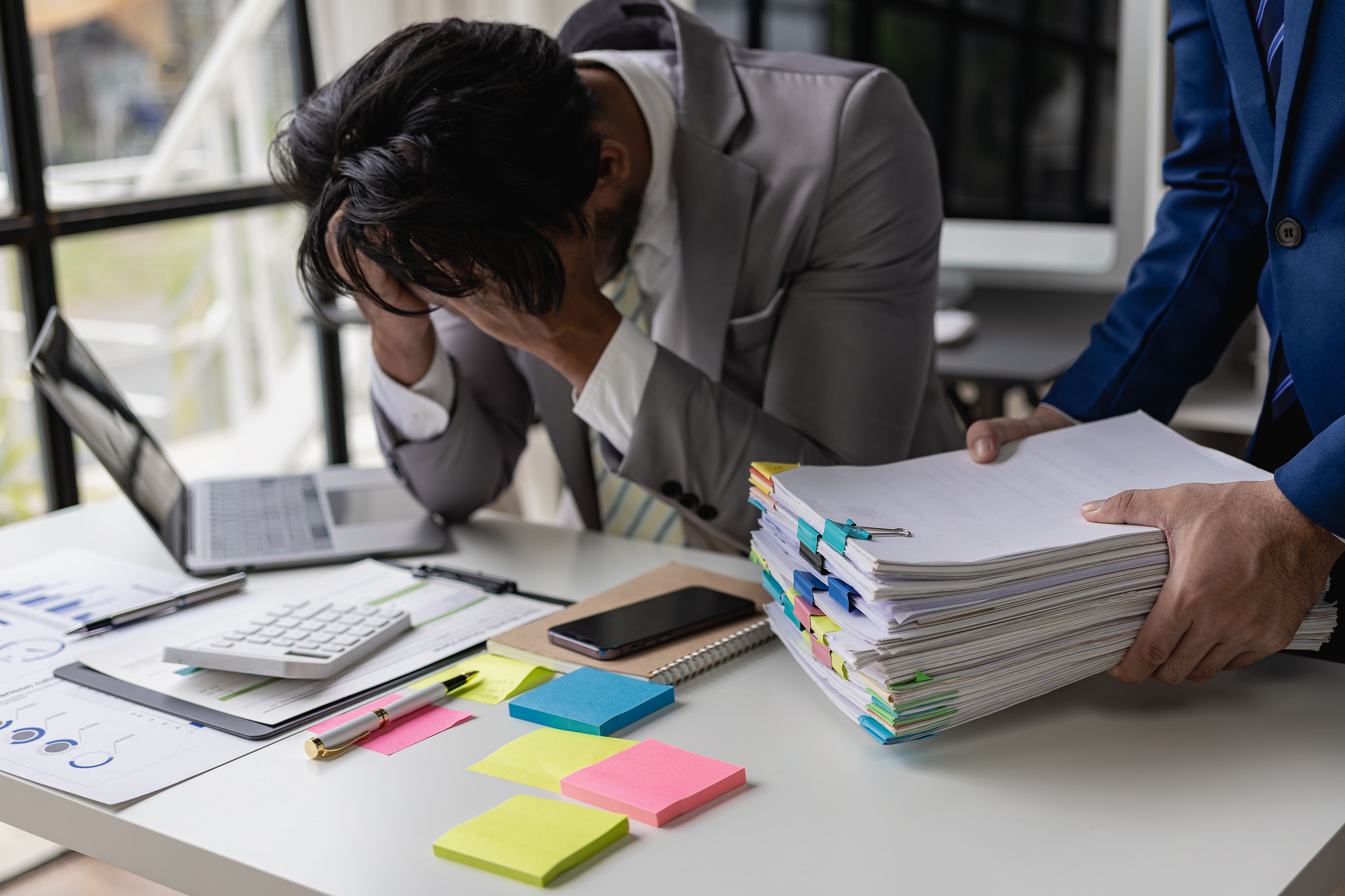Male accountant stressed with too much paperwork and exhausted from work, covering his face with friend holding a lot of documents.