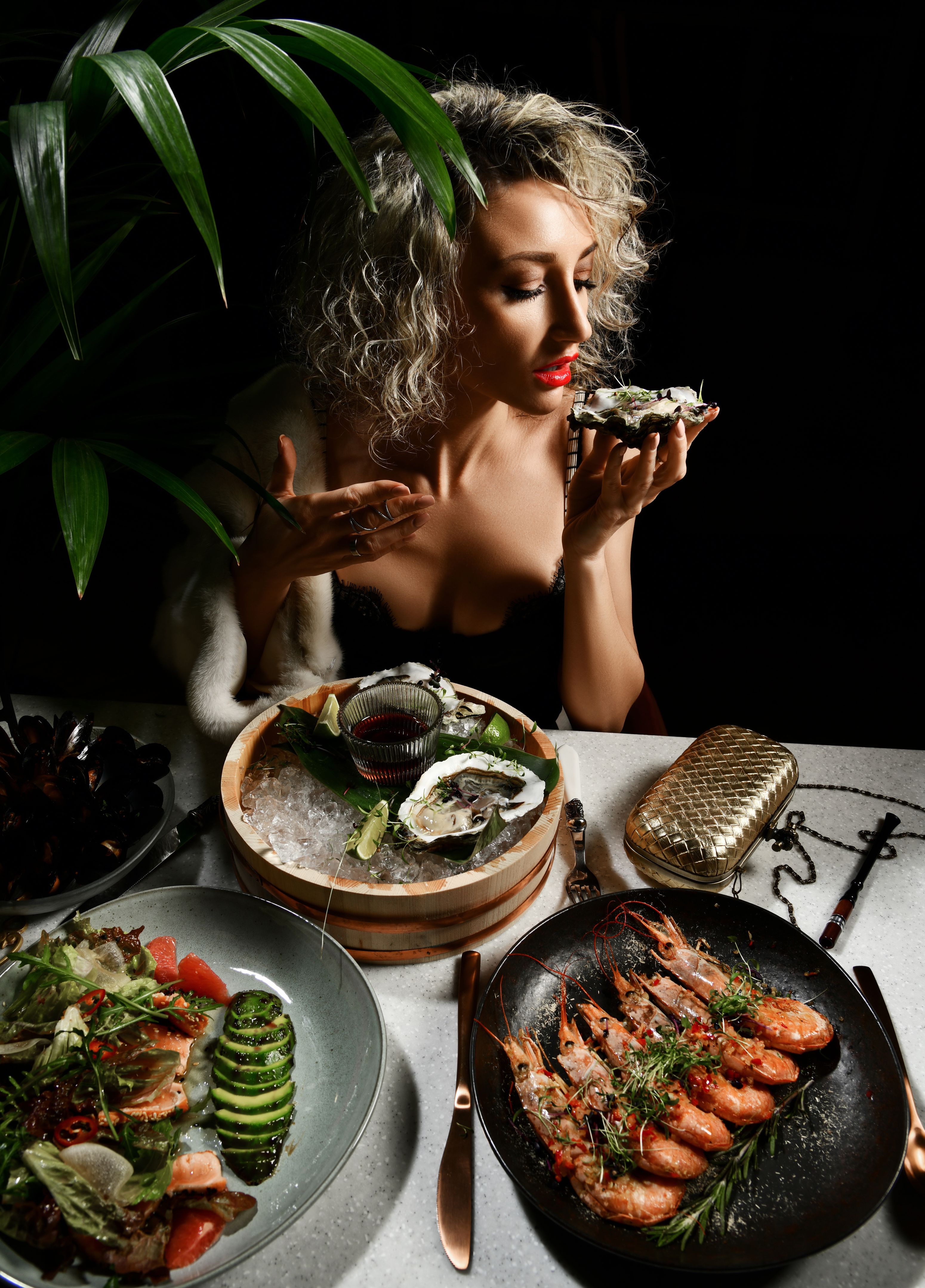 Blonde curly young woman in expensive evening dress eats oysters, prawns, mussels and salad in seafood restaurant Blonde curly young woman in expensive evening dress eats oysters, prawns, mussels and salad in seafood restaurant
