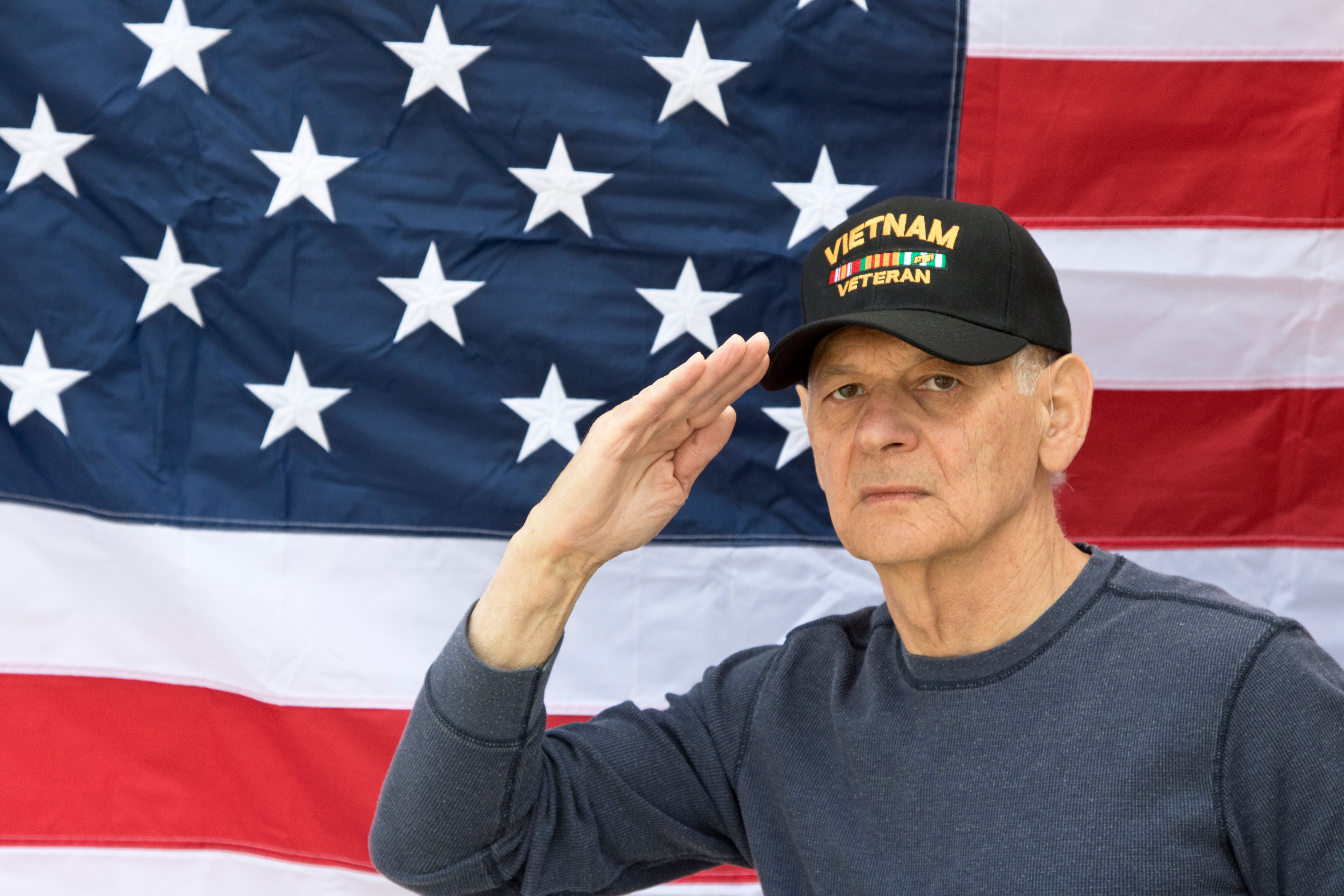 Vietnam Veteran Saluting looking angry wearing Veterans cap, with American Flag in background.