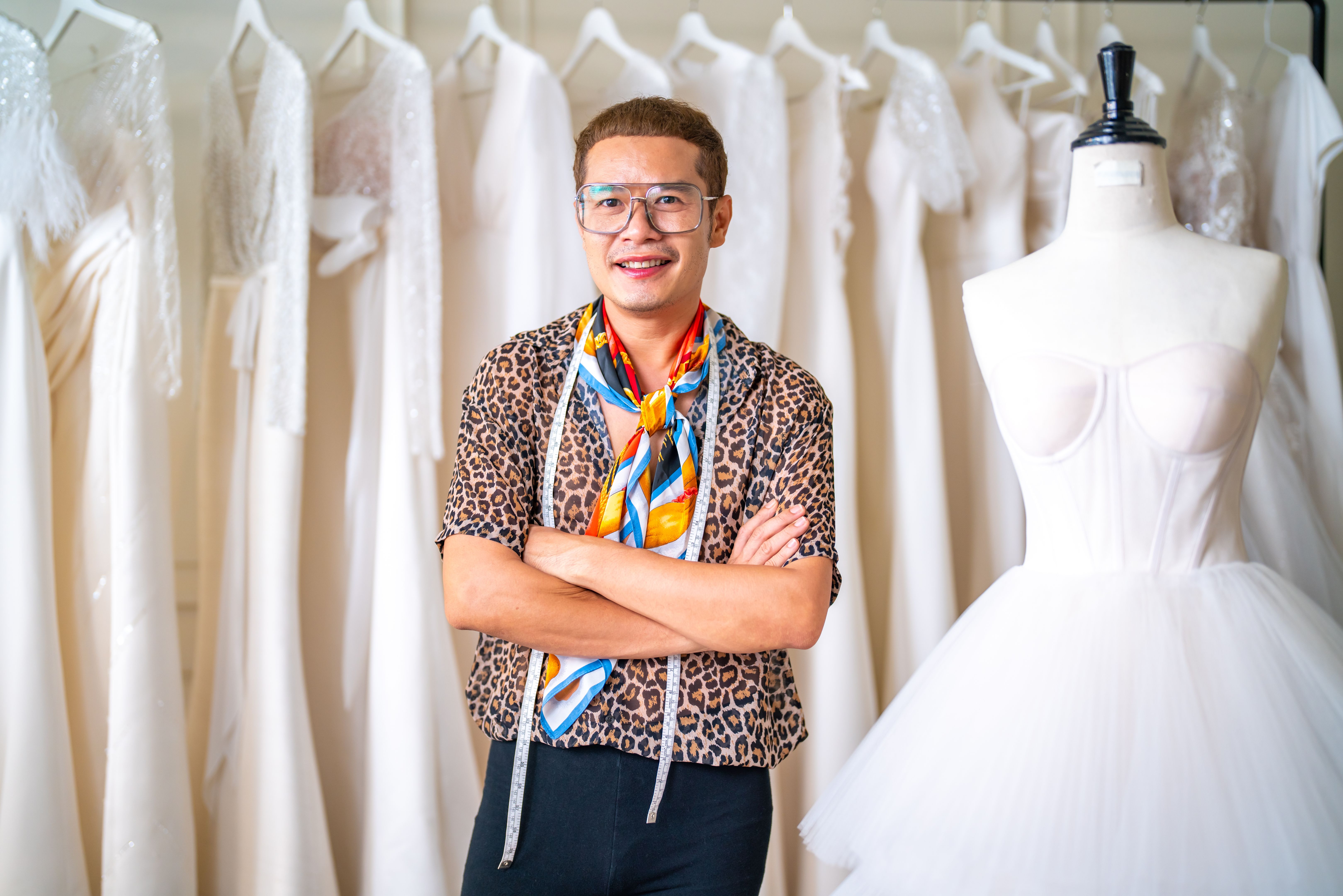 Portrait of Asian LGBTQ guy designer using tape measure measuring wedding dress on sewing mannequin