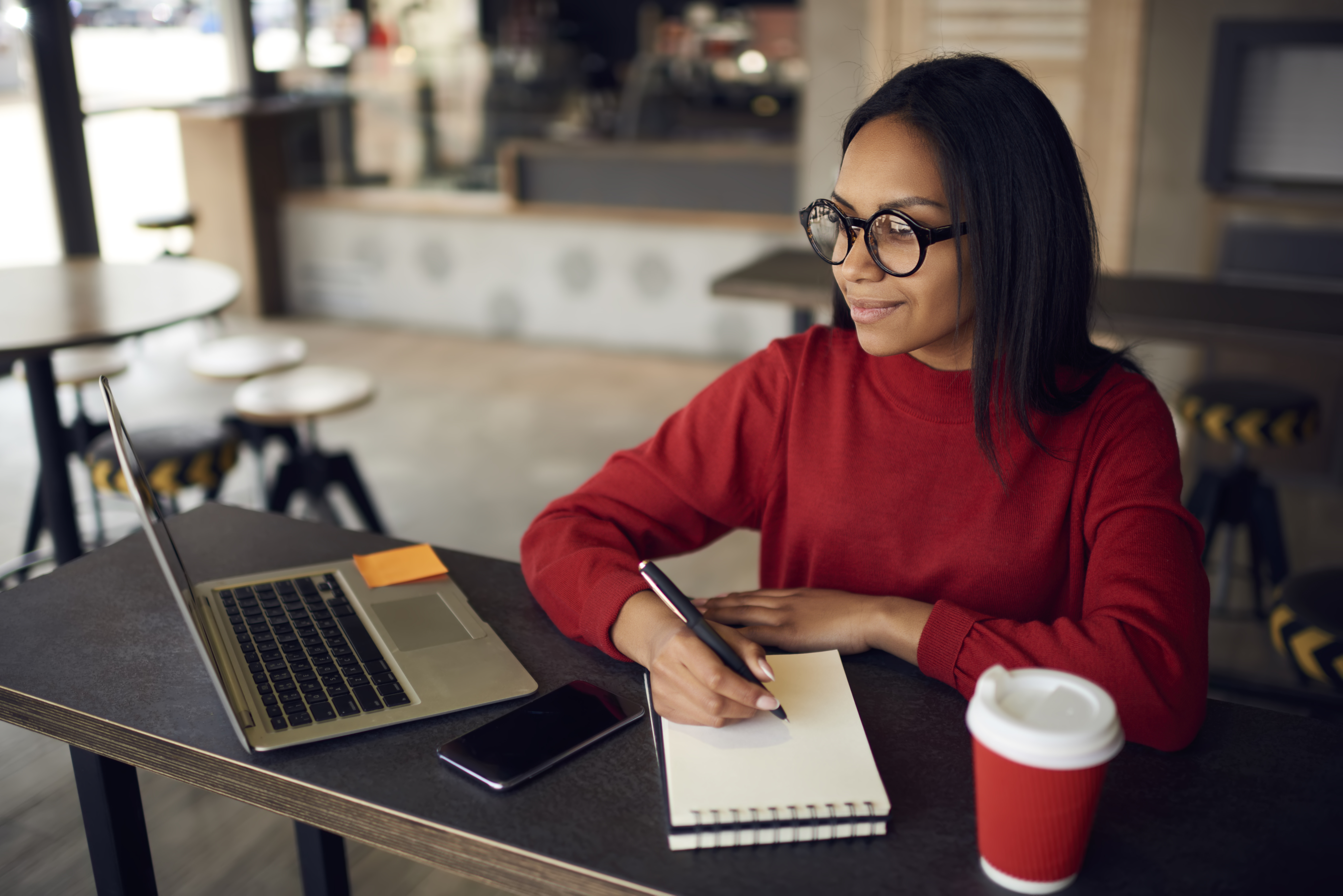 Positive dark skinned hipster girl in optical spectacles for vision correction looking away while creating new article with romantic idea in textbook for posting at own web blog via laptop device