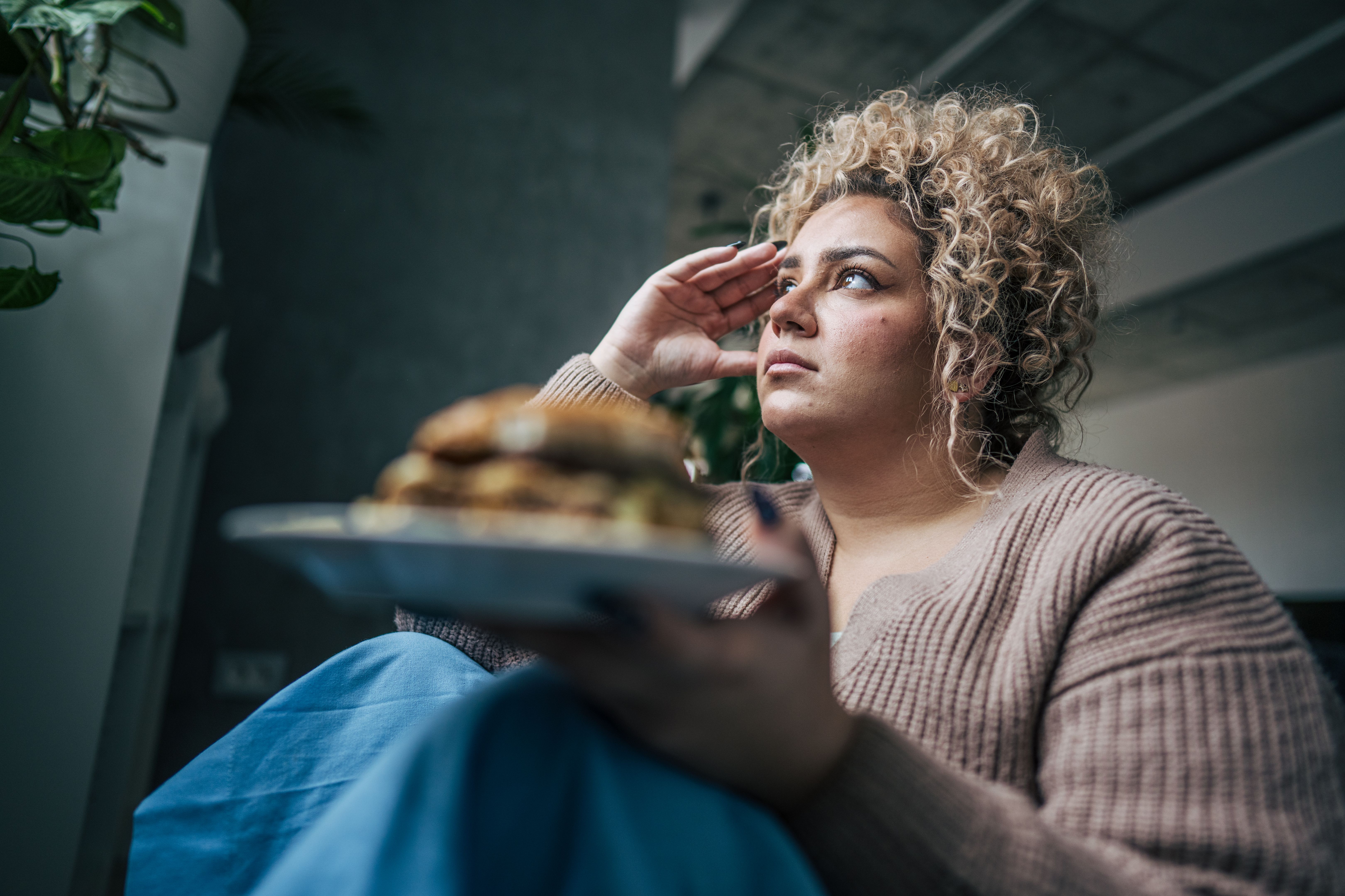 Chubby woman sitting beside the bed in a quiet bedroom, she holds a plate with burger and crispy fries, but her expression is far from enjoyment. Chubby woman sitting beside the bed in a quiet bedroom, she holds a plate with burger and crispy fries, but her expression is far from enjoyment.
