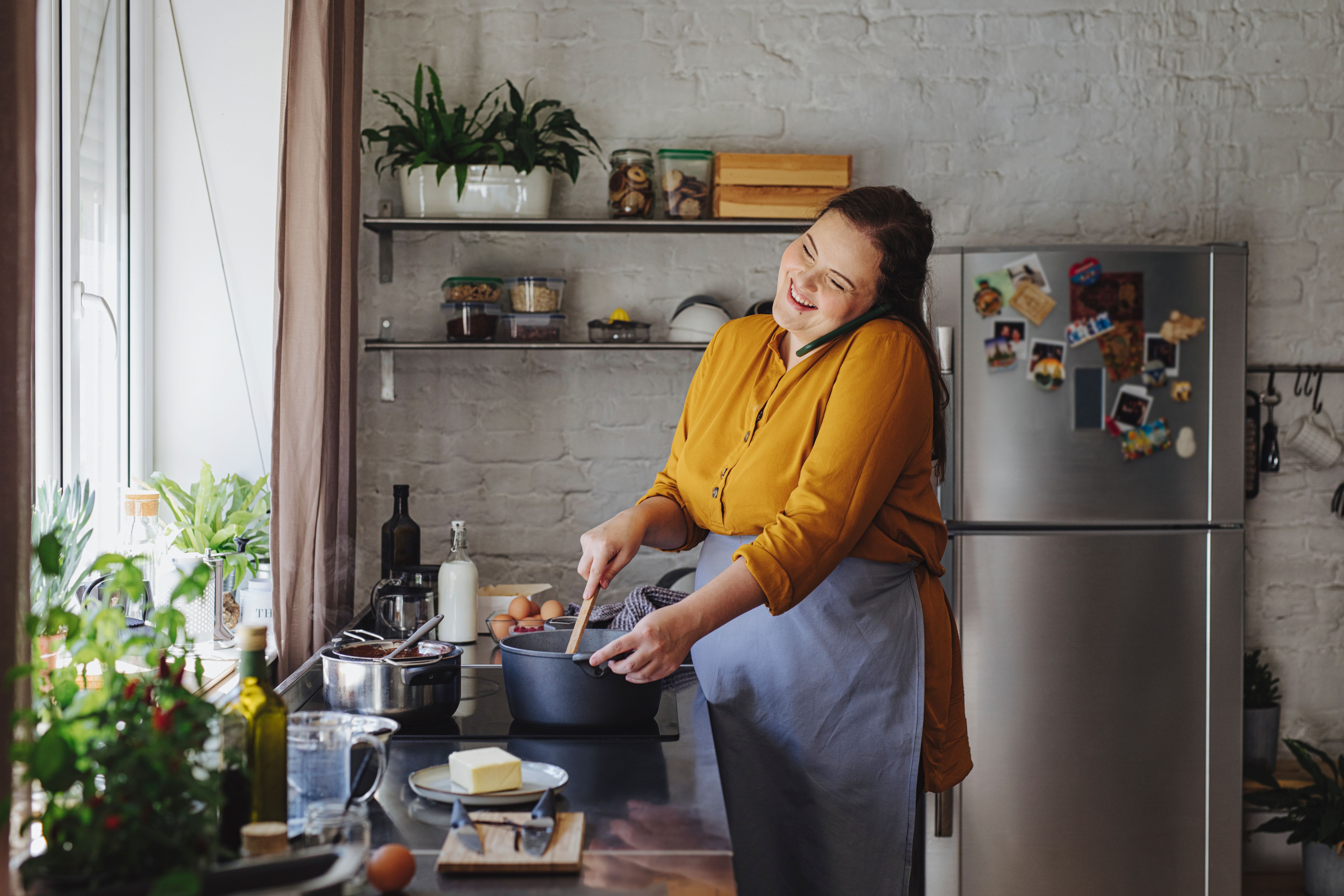 Beautiful Young Cheerful Woman Having a Phone Call while Mixing Hot Sauce in a Pan over a Kitchen Stove