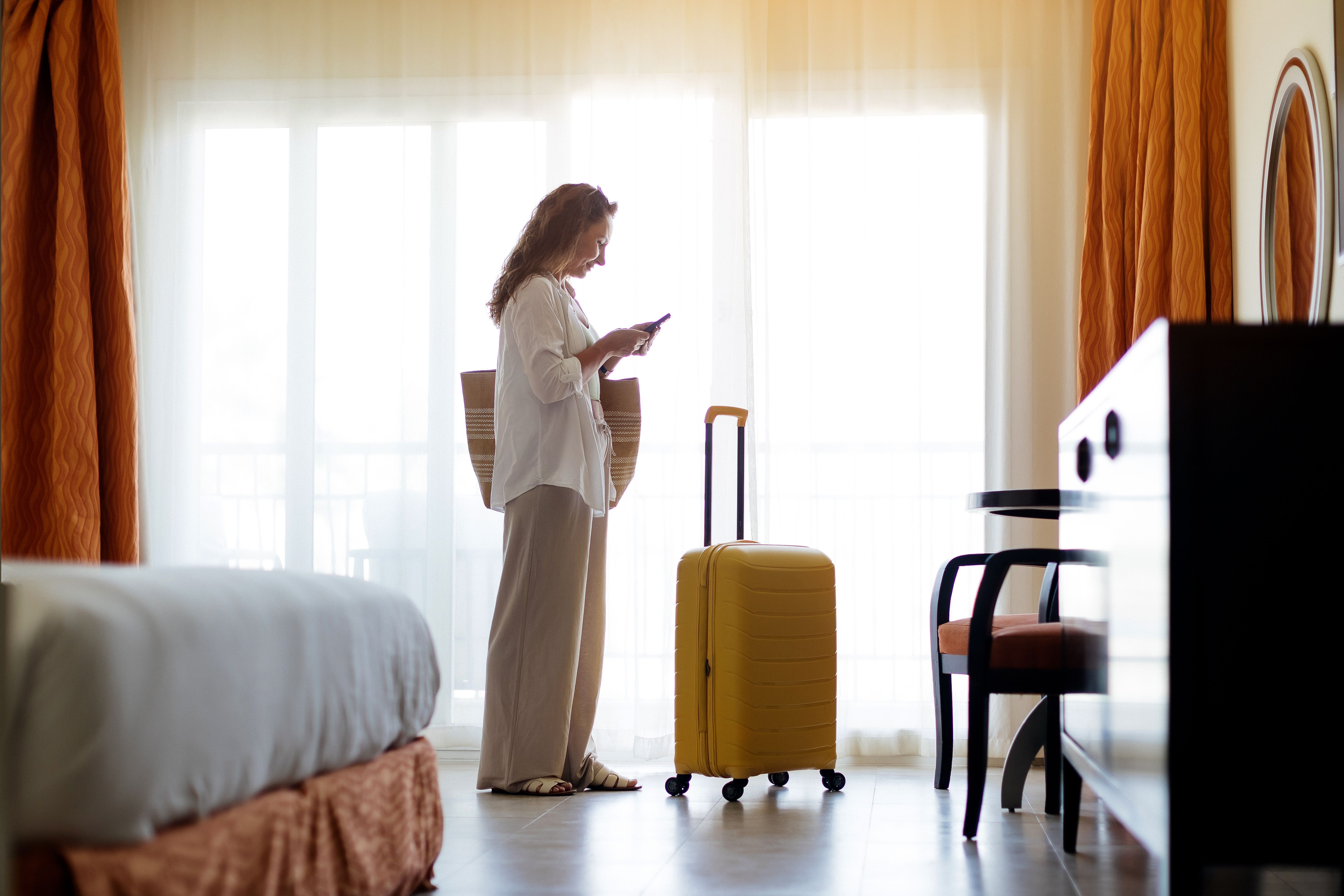 Woman standing in hotel room with yellow suitcase, looking at her phone Woman standing in hotel room with yellow suitcase, looking at her phone