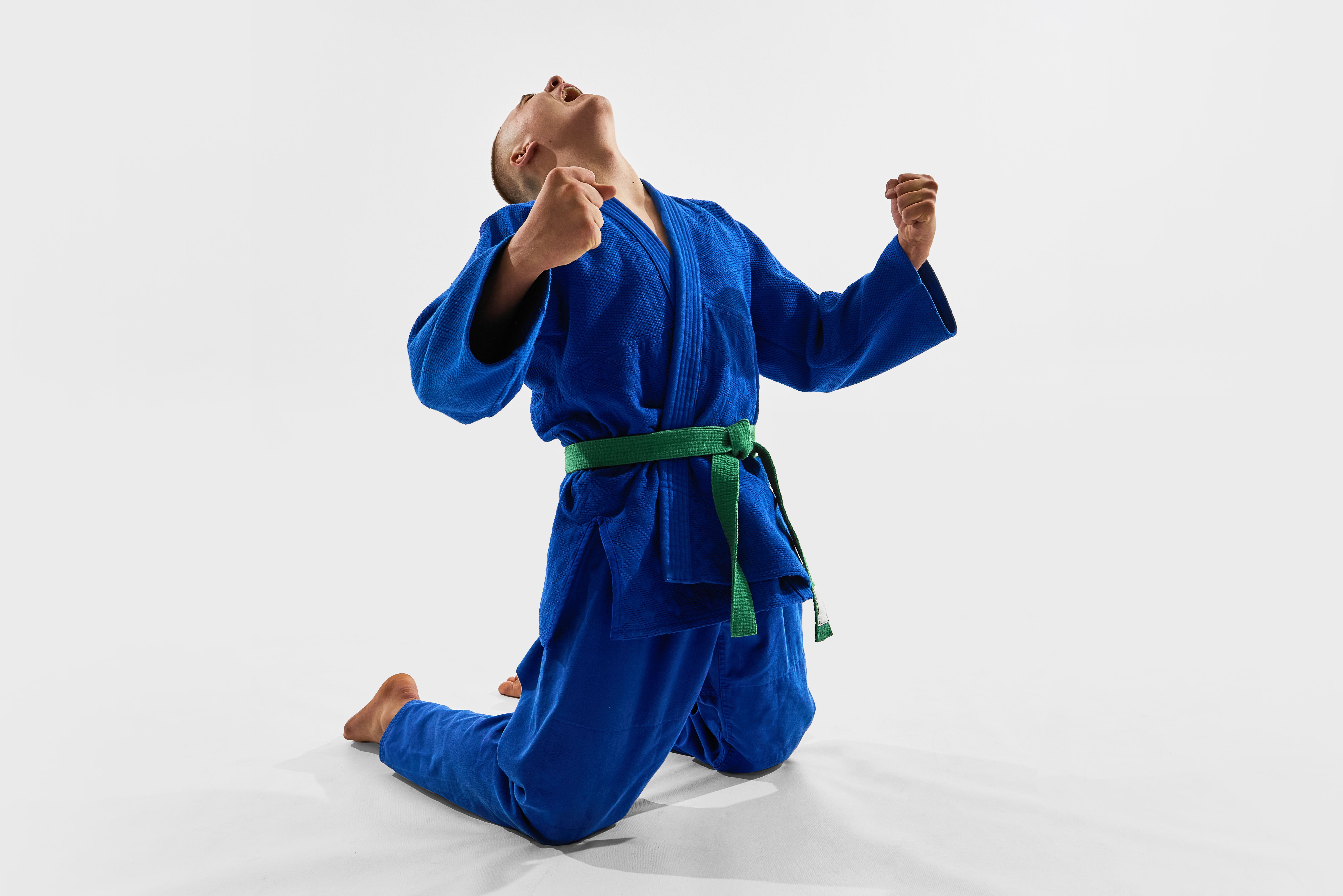 Triumphant athlete in blue gi kneels with raised fists and open mouth, symbolizing raw emotion, personal strength, and victorious spirit of combat championship. Triumphant athlete in blue gi kneels with raised fists and open mouth, symbolizing raw emotion, personal strength, and victorious spirit of combat championship.