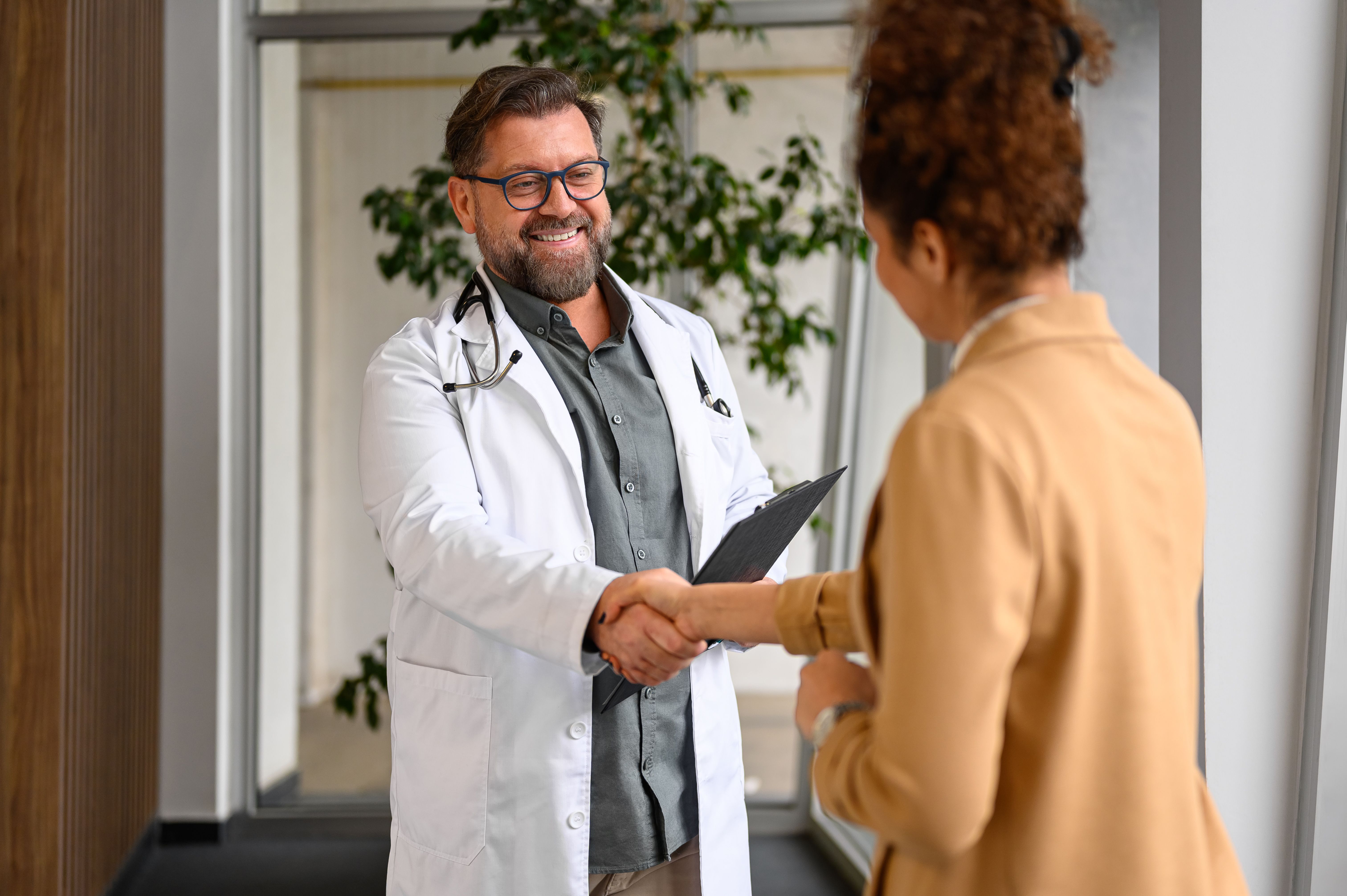 Positive male doctor in lab coat and glasses shaking hands with young female patient in modern hospital