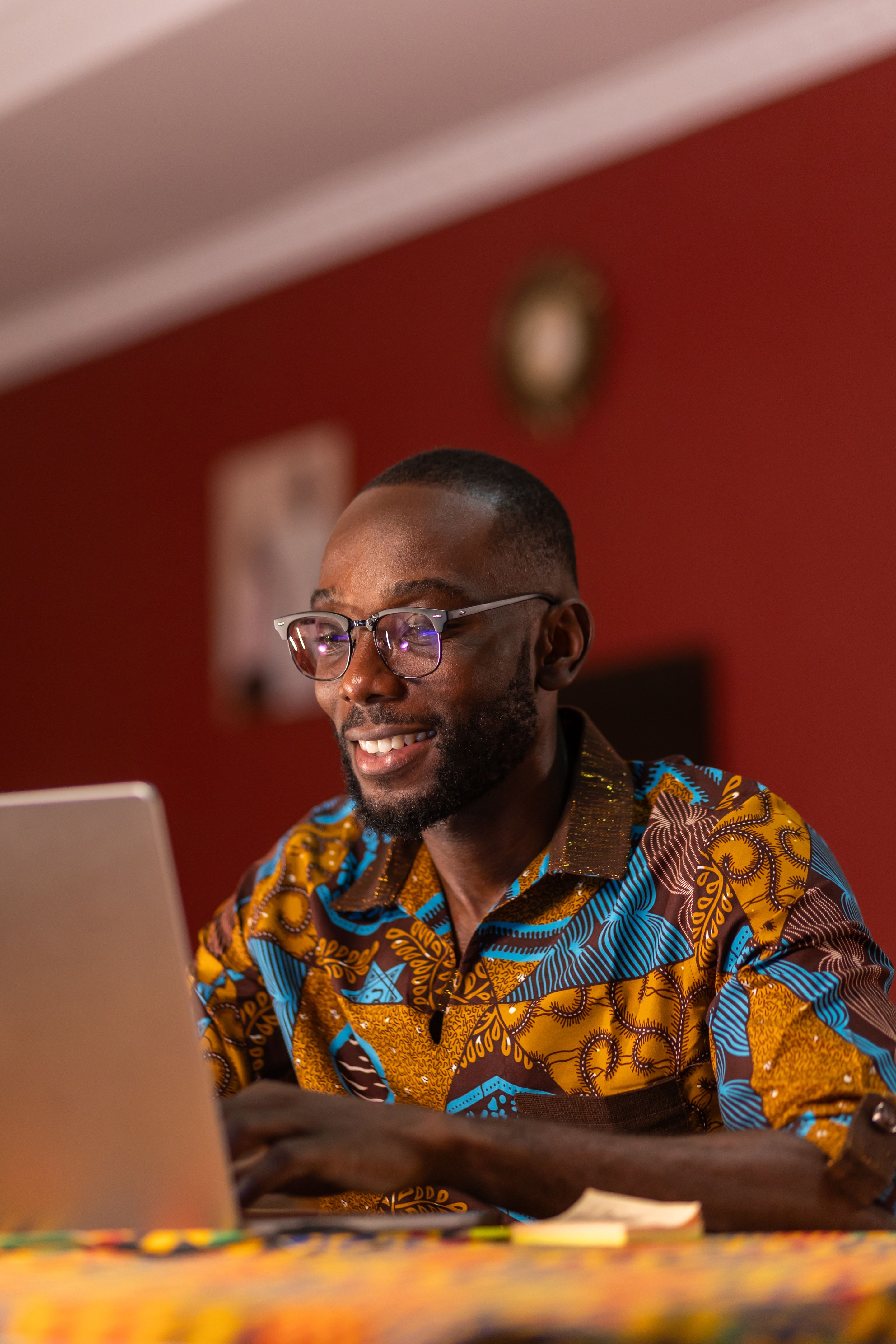 A young African man in a colorful shirt works on his laptop in a dimly lit room.