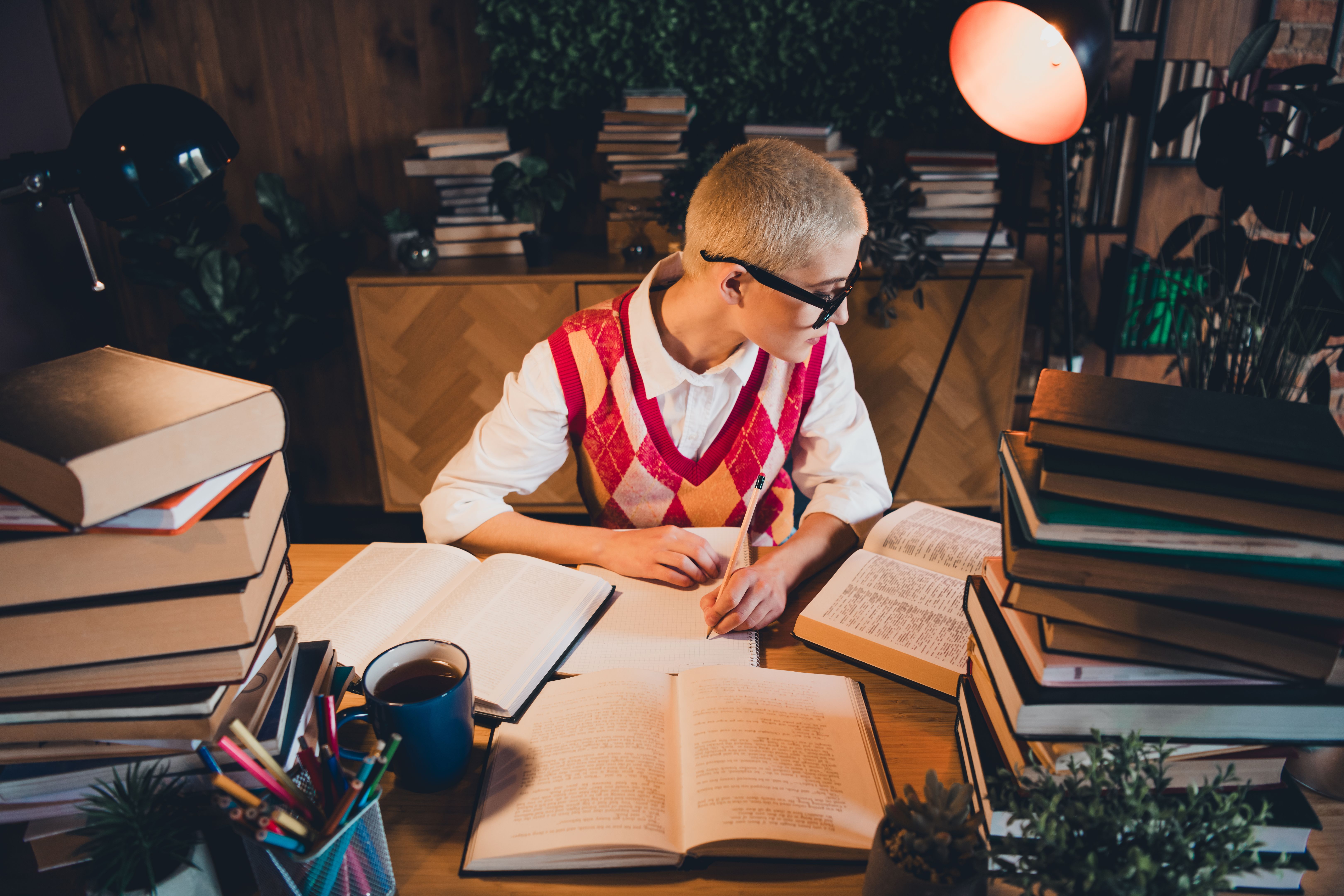 Young woman with short blond hair wearing glasses and a stylish argyle waistcoat studies late in her cozy apartment