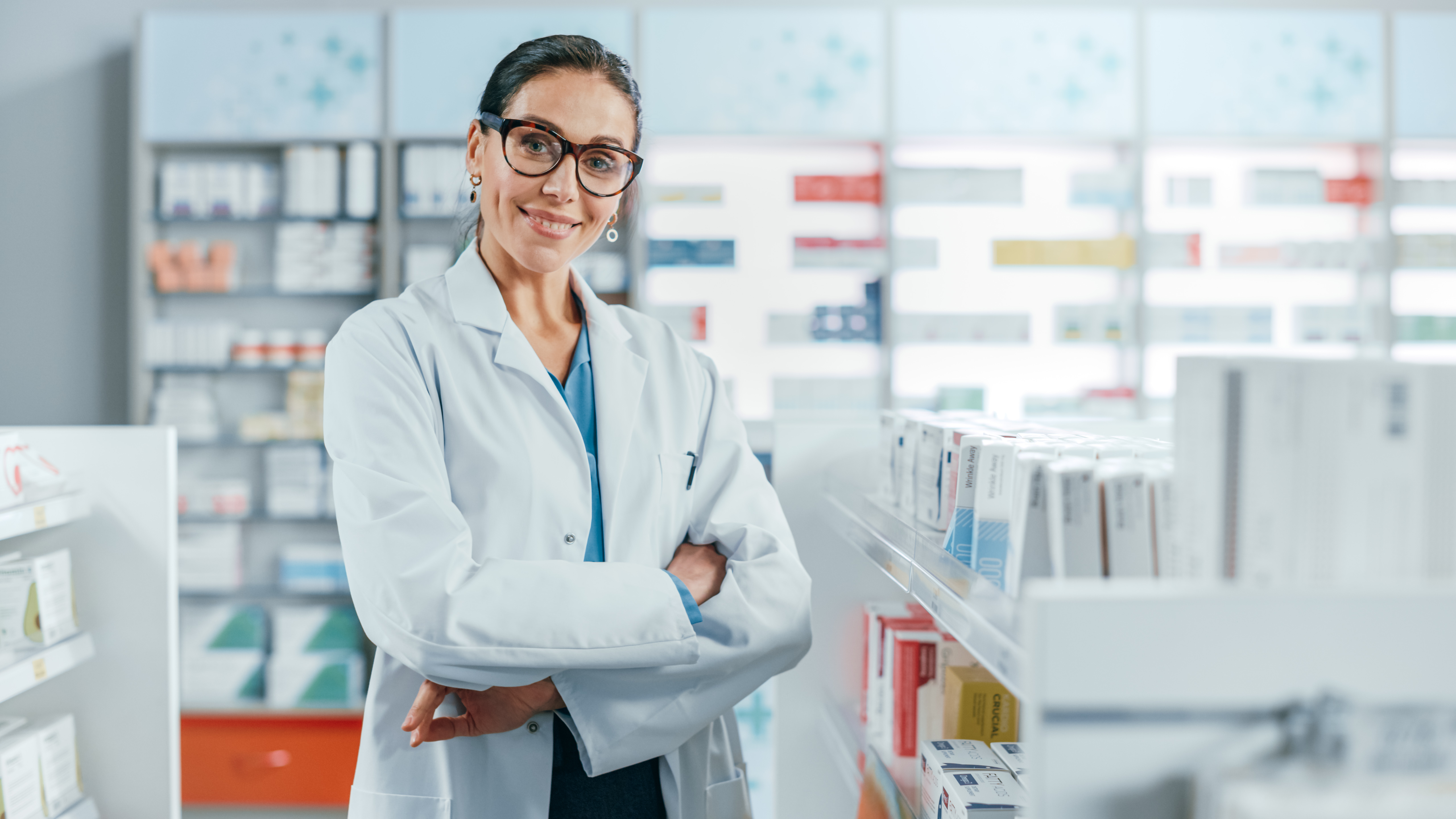 Pharmacy: Portrait of Beautiful Professional Caucasian Female Pharmacist Wearing Glasses, Crosses Arms and Looks at Camera Smiling Charmingly. Drugstore Store with Shelves Health Care Products