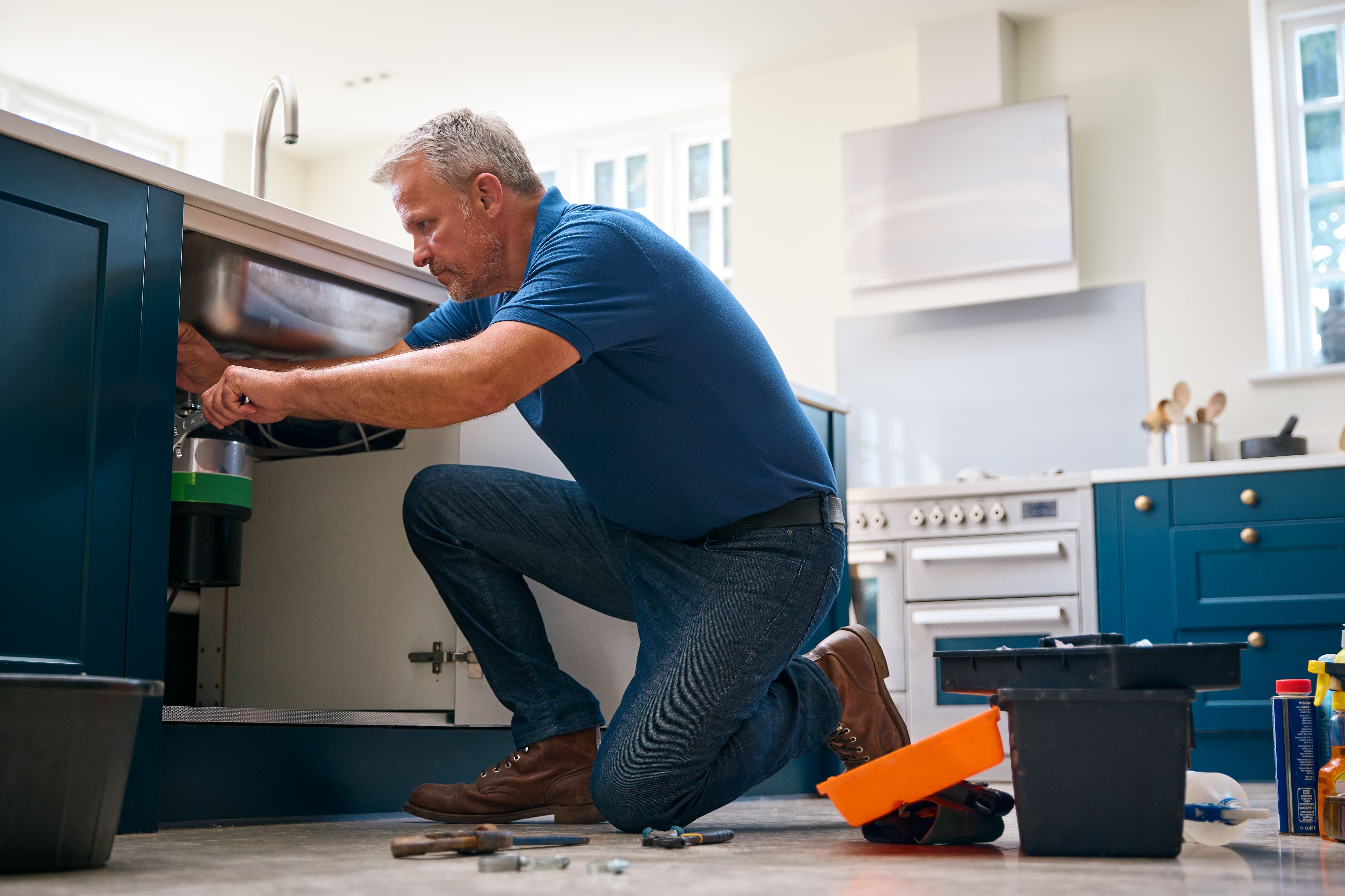 Mature Male Plumber Fixing Waste Disposal Unit In Domestic Kitchen Sink