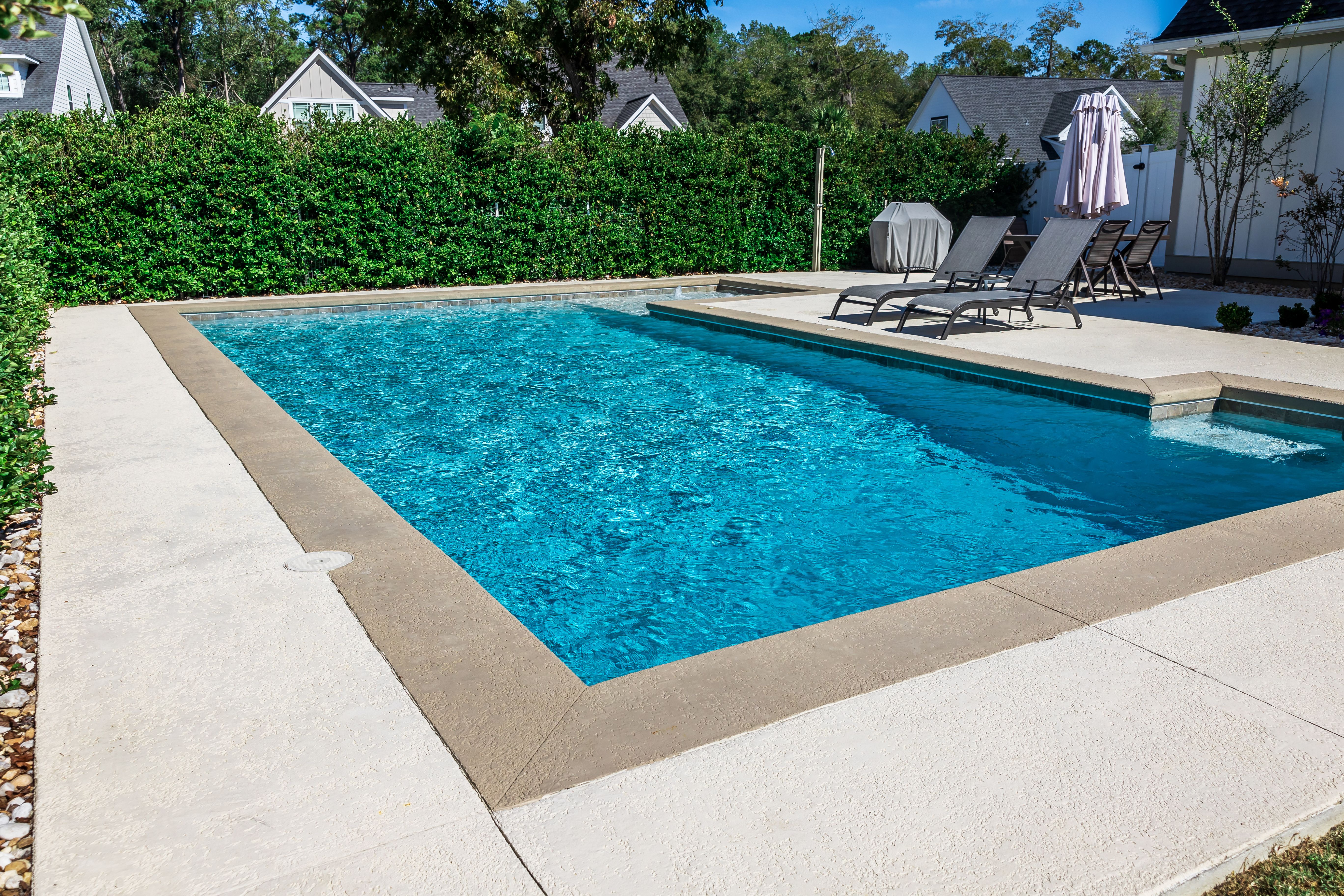 A rectangular new swimming pool with tan concrete edges in the fenced backyard of a new construction house