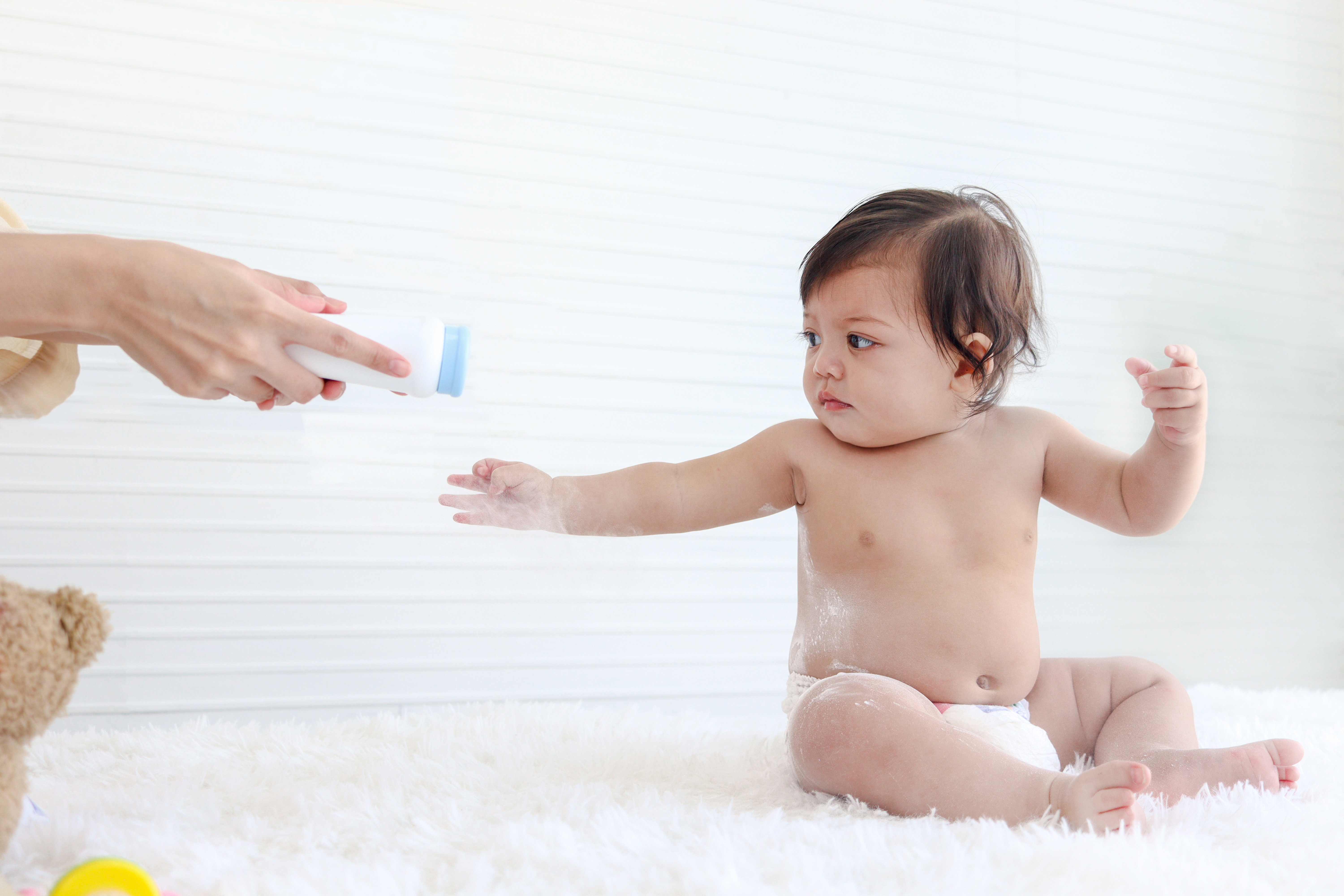 Portrait of happy smiling toddle baby girl sit on fluffy white rug with mother hand hold dusting powder bottle, mom apply talcum powder on body of little cute kid daughter child, childhood care family Portrait of happy smiling toddle baby girl sit on fluffy white rug with mother hand hold dusting powder bottle, mom apply talcum powder on body of little cute kid daughter child, childhood care family