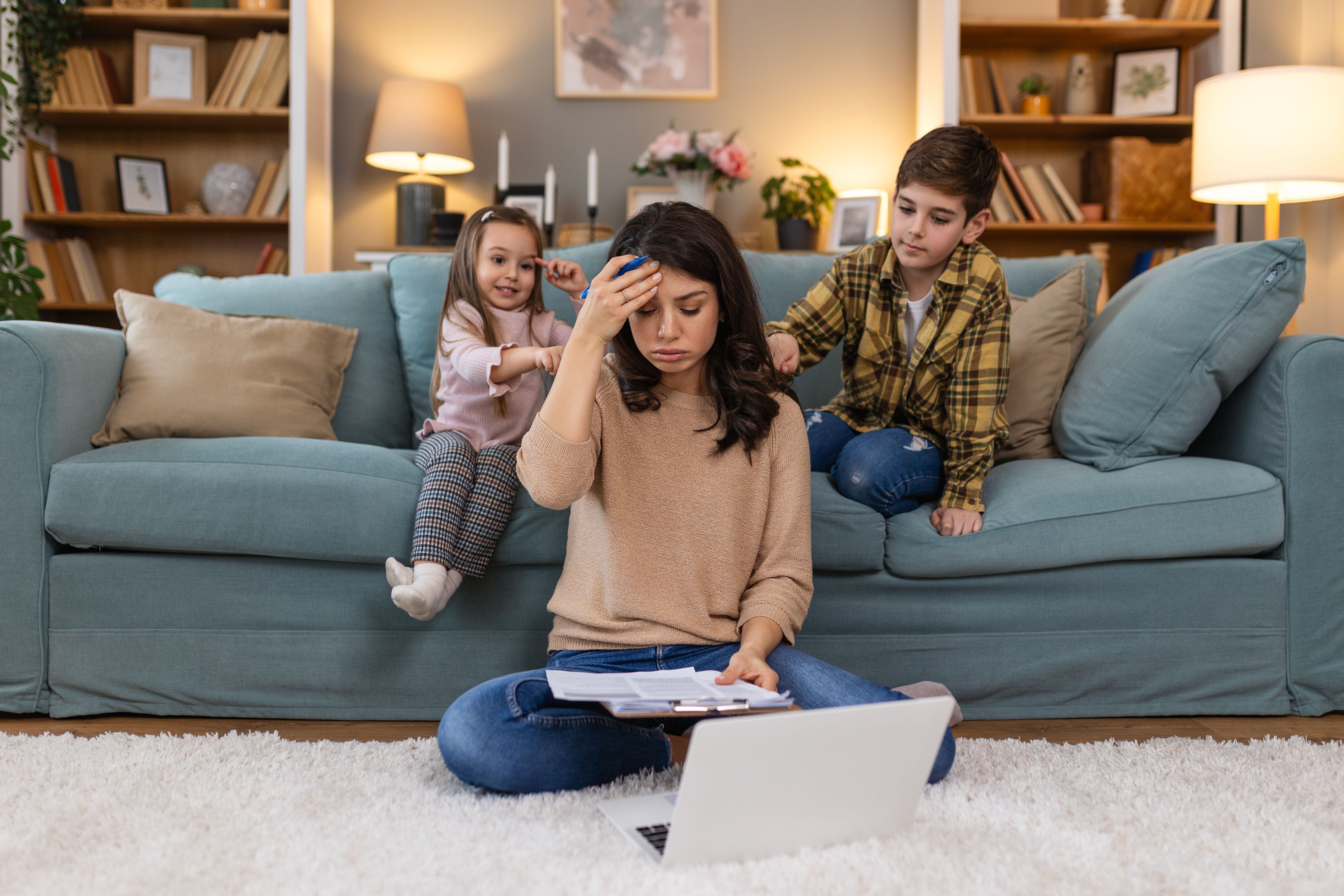 Tired young mother sitting on the floor and working with laptop and documents while little kids and jumping on the sofa and having fun and making noise