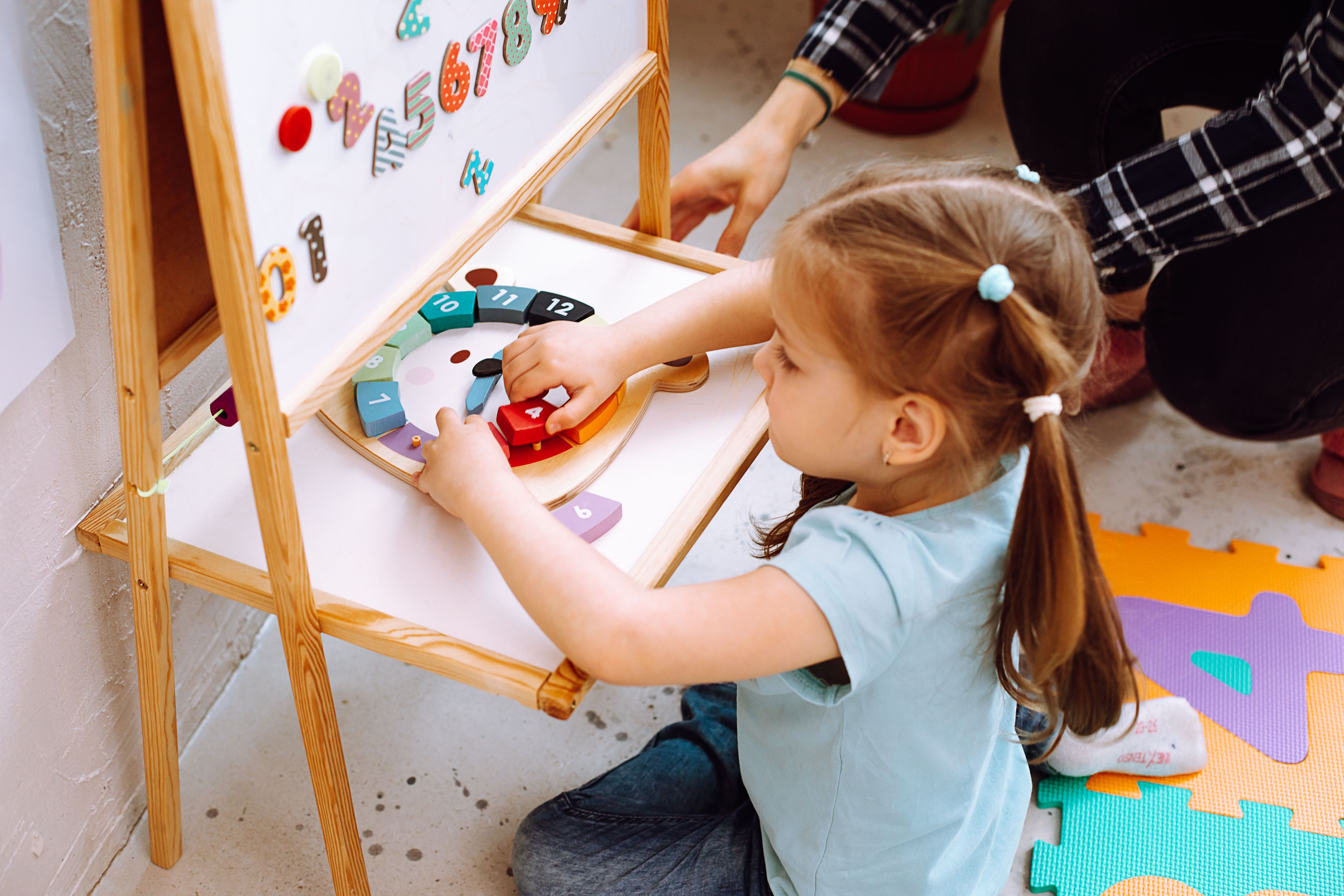 Side view of nice little girl sitting at magnetic board, playing with numbers of clock with help of teacher woman. Side view of nice little girl sitting at magnetic board, playing with numbers of clock with help of teacher woman.