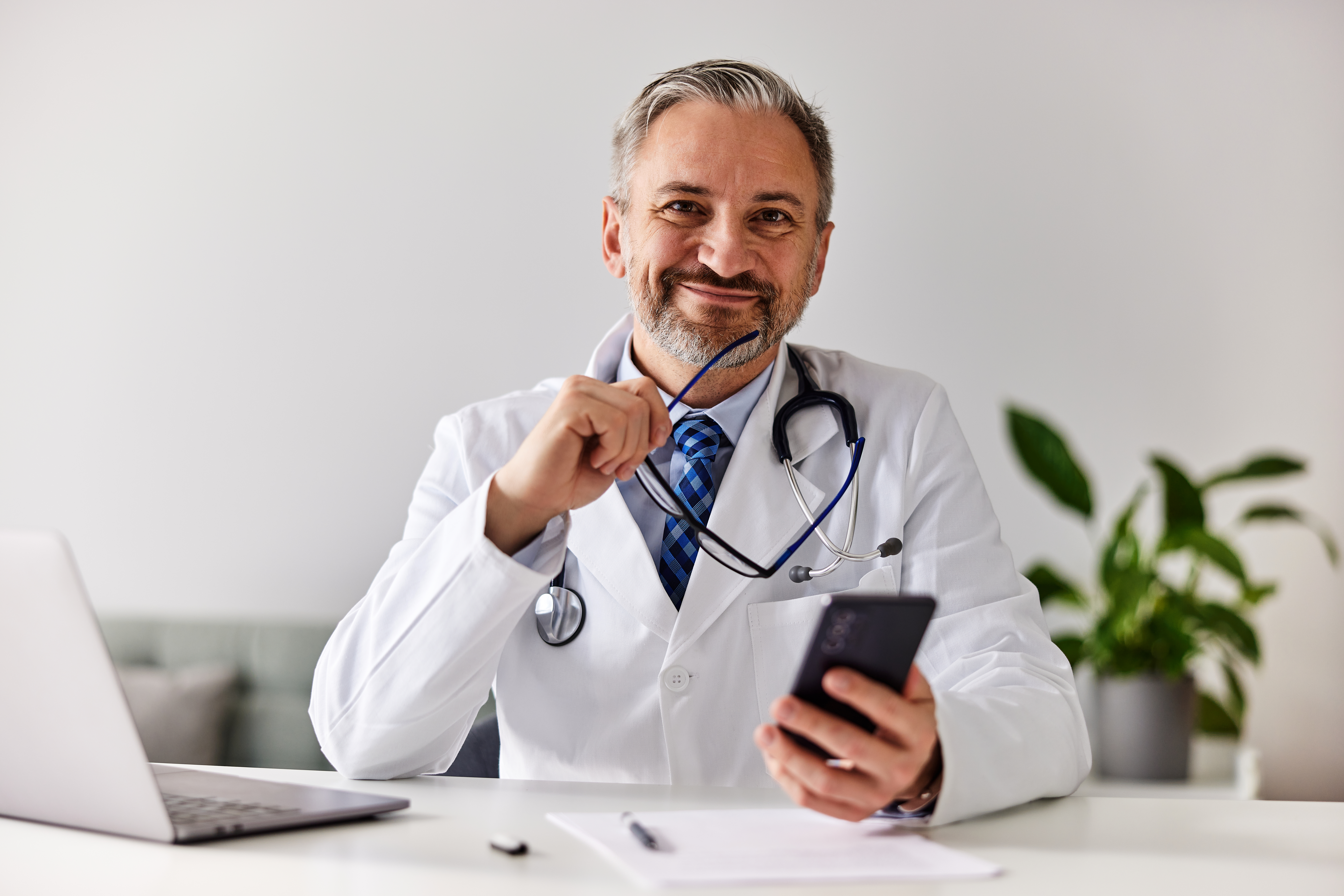 Portrait of a smiling doctor holding glasses and a mobile phone at the office.