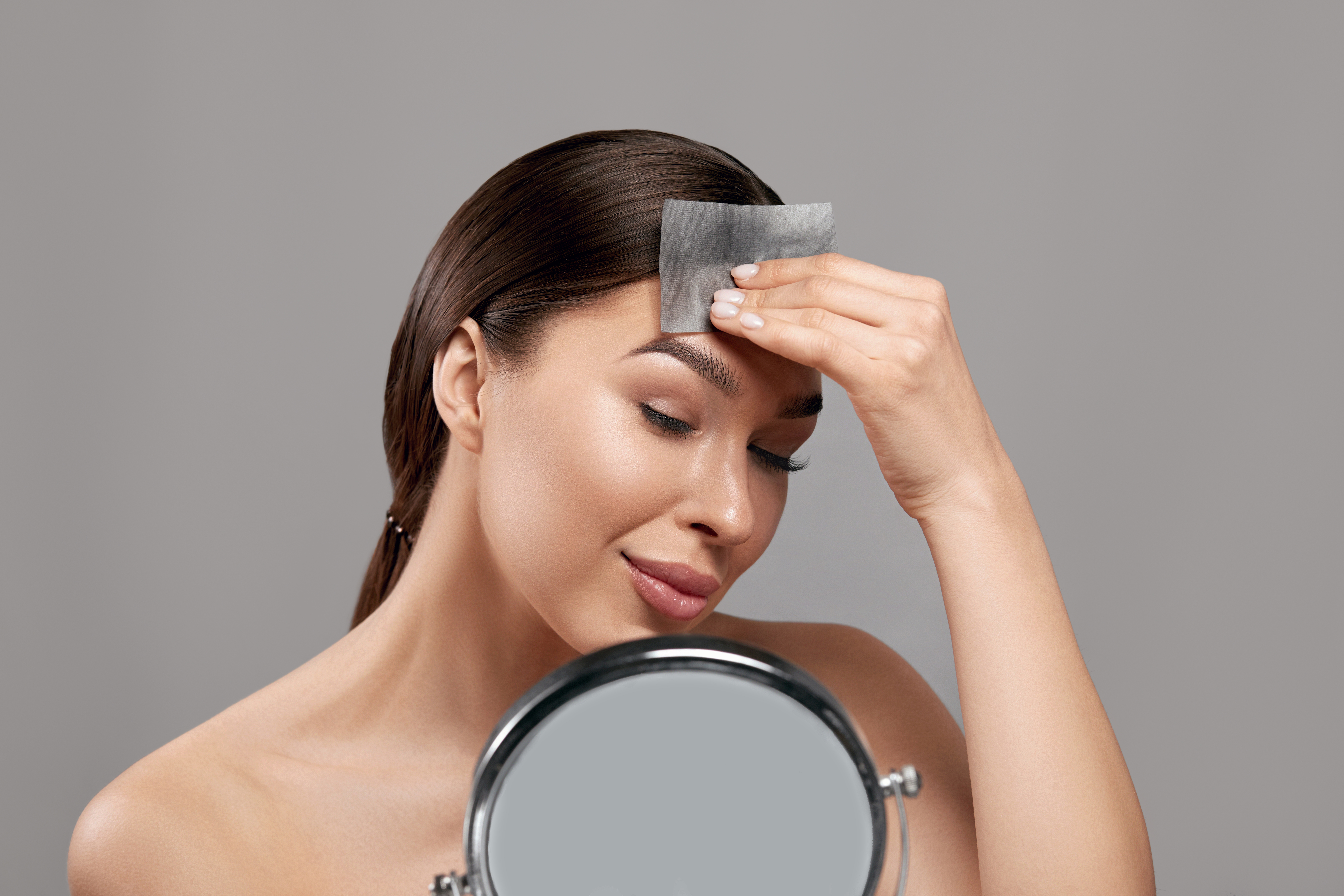Beautiful woman using facial oil blotting paper. Photo of woman with perfect makeup on gray background. Beauty concept. Cosmetology, cosmetics. Facial treatment