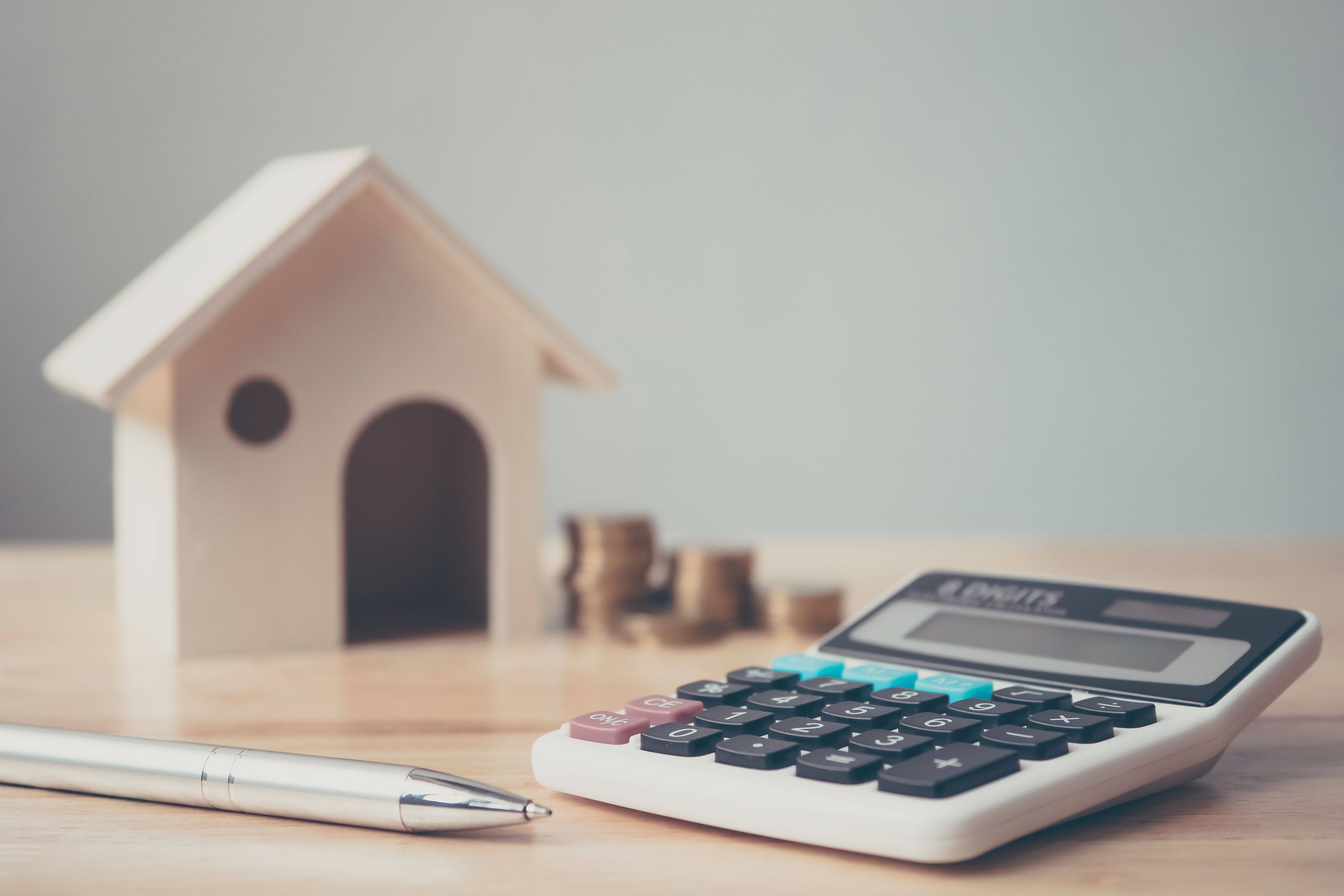 Calculator with wooden house and coins stack and pen on wood table. Property investment and house mortgage financial concept Calculator with wooden house and coins stack and pen on wood table. Property investment and house mortgage financial concept
