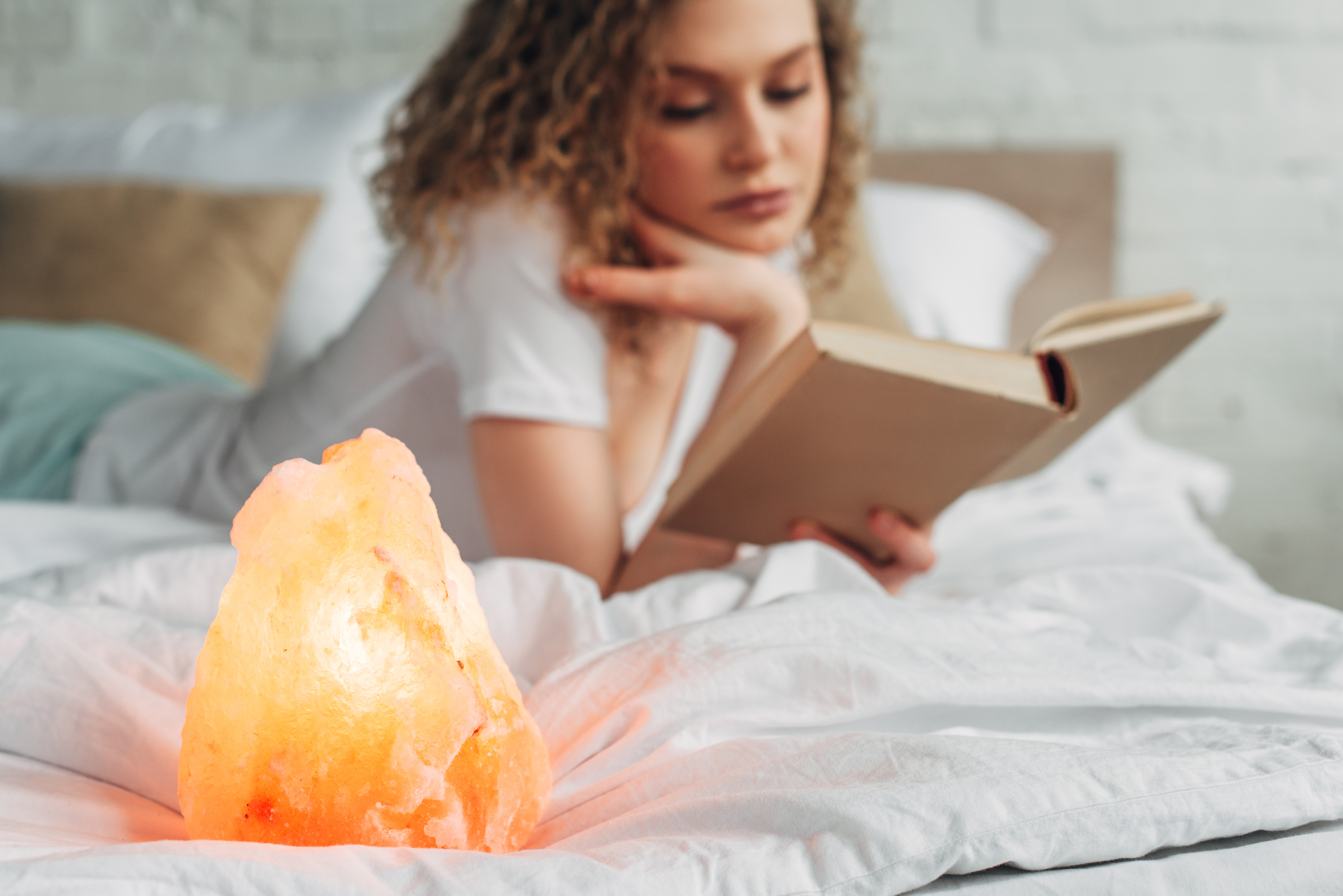 selective focus of curly woman reading book on bed with Himalayan salt lamp