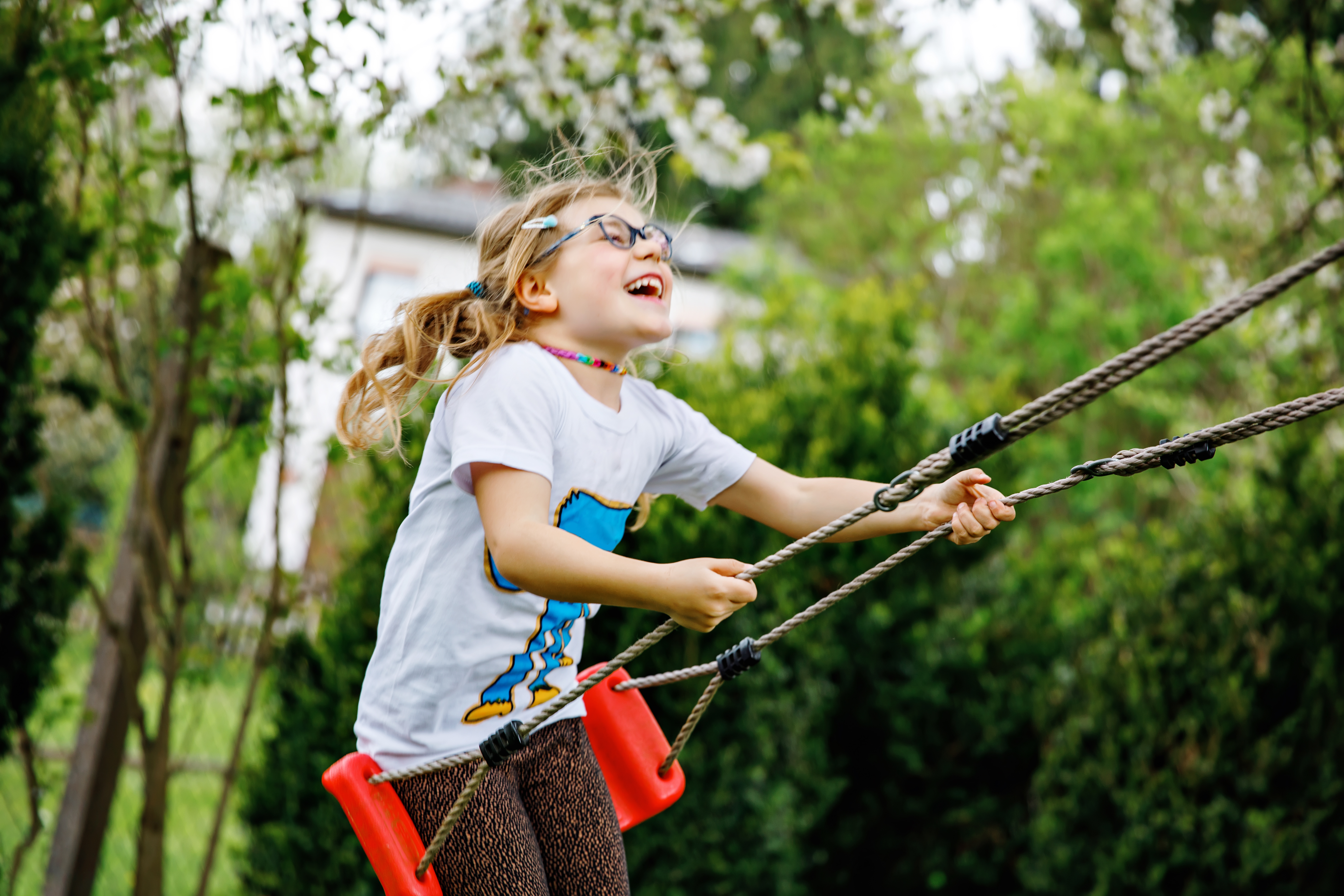 Happy little preschool girl with glasses having fun on swing in domestic garden. Cute healthy child swinging under blooming trees on sunny spring day. Kid laughing and crying.