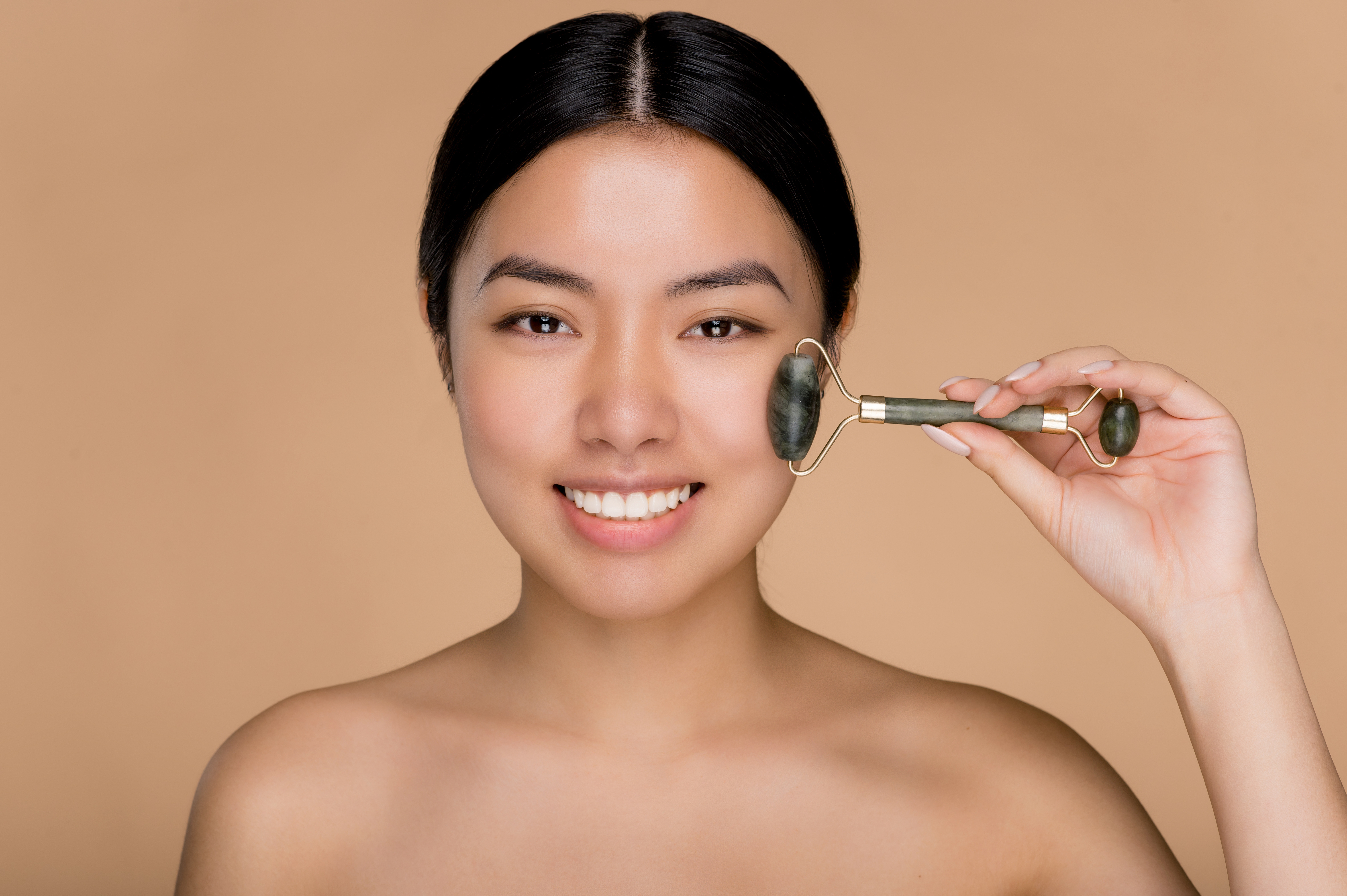 Close up photo of charming beautiful young Asian woman with naked shoulders, using derma roller for massage her facial skin. Photo of a smiling woman on isolated beige background