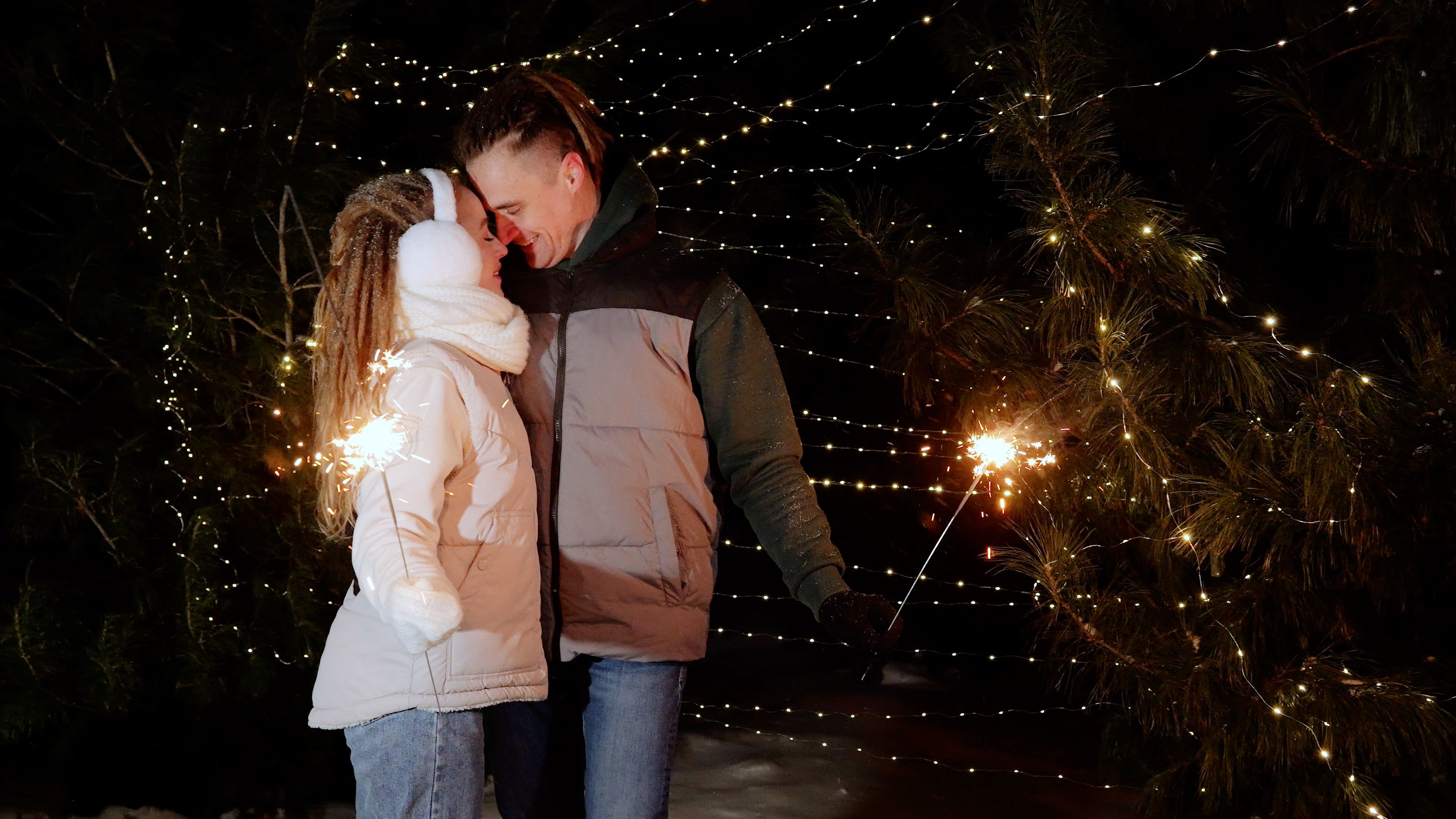 Couple holding sparkling fireworks, celebrating festive night amid illuminated winter forest with glowing holiday lights