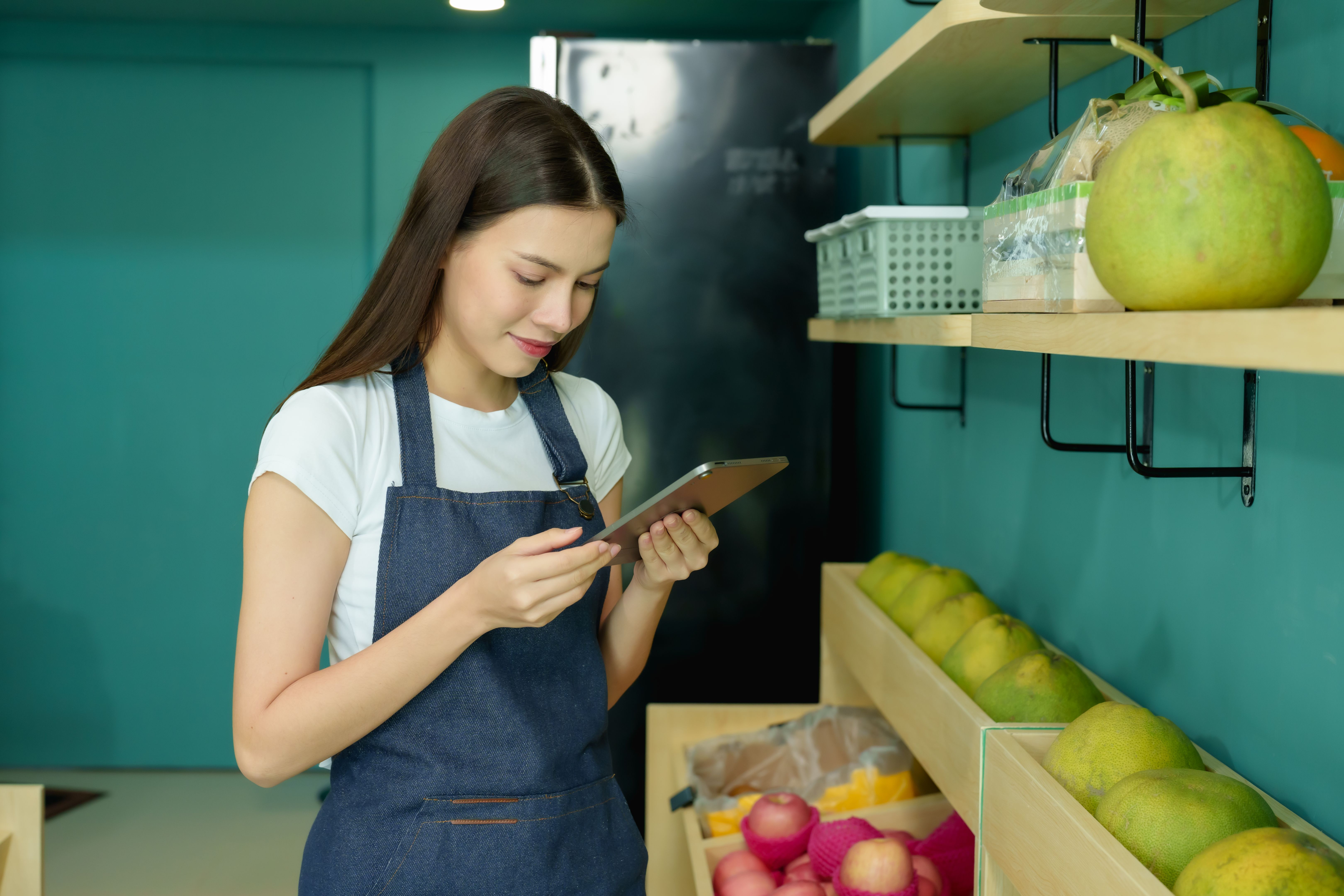 Confident female shopkeeper holding a ripe mango and using a tablet for inventory in her small tropical fruit store. Reflecting efficiency and innovation in small business management. Confident female shopkeeper holding a ripe mango and using a tablet for inventory in her small tropical fruit store. Reflecting efficiency and innovation in small business management.