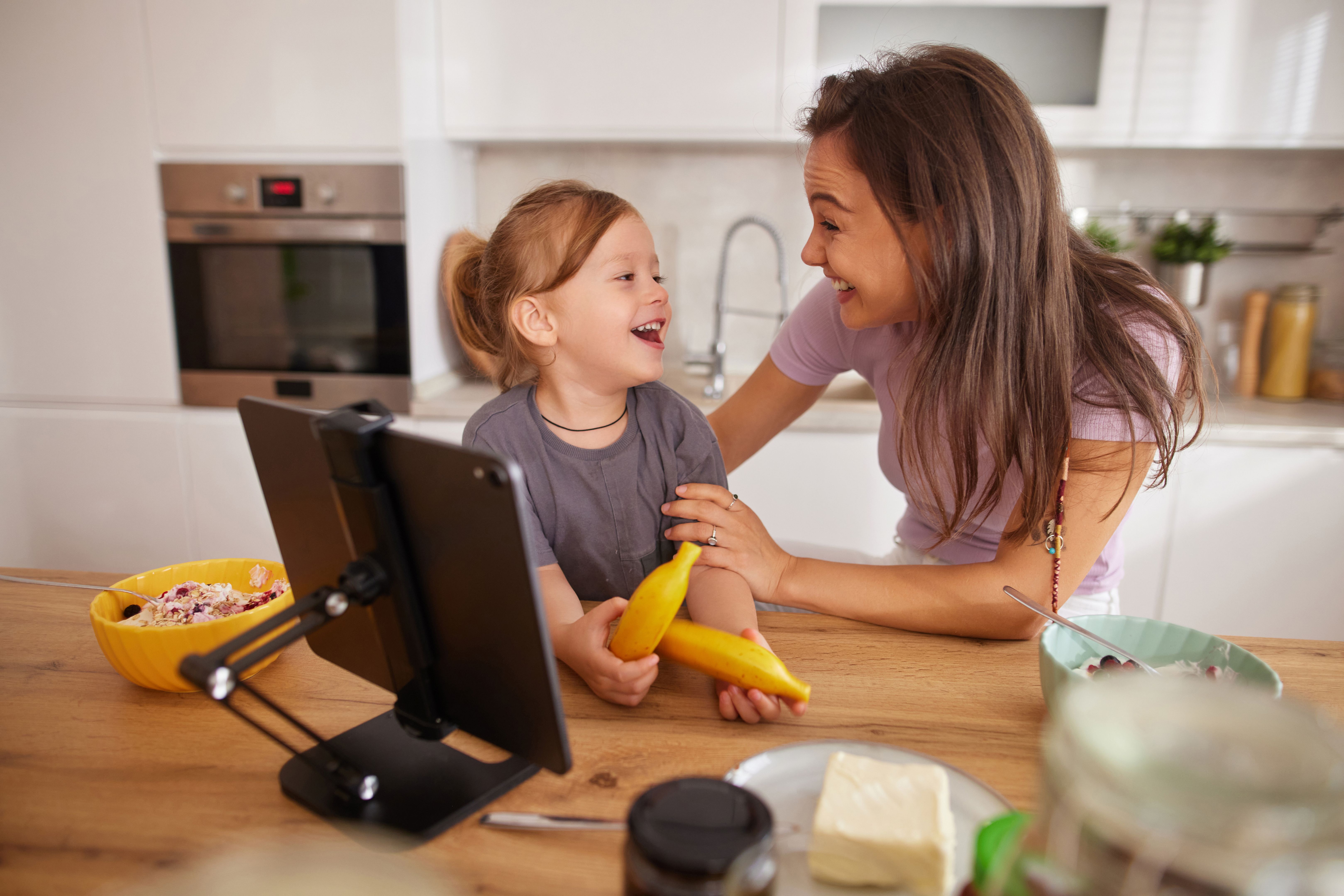 Mother and daughter laughing, using tablet during breakfast in kitchen