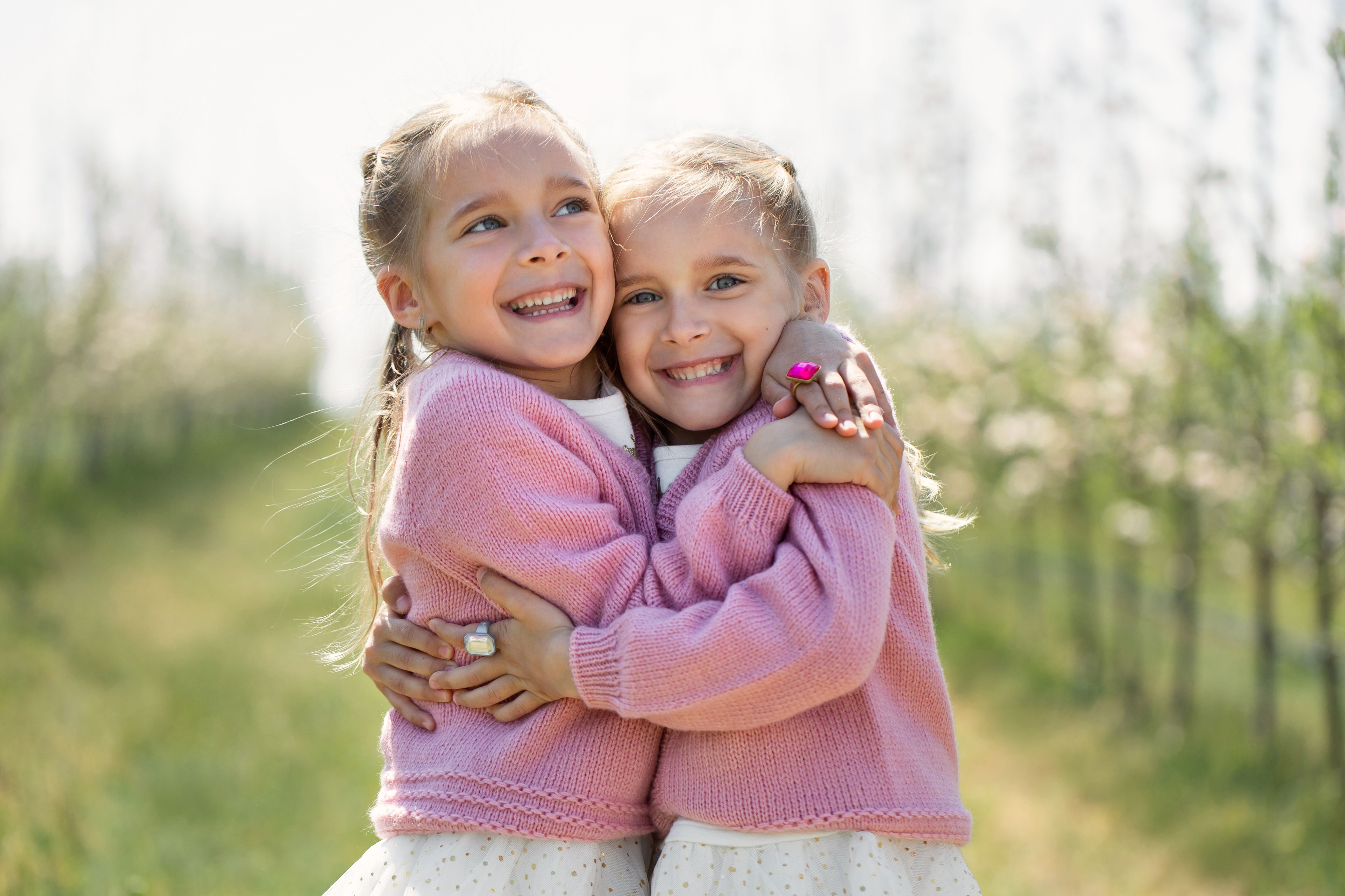 Happy two twin sisters are hugging against the background of a green blossoming apple orchard Happy two twin sisters are hugging against the background of a green blossoming apple orchard