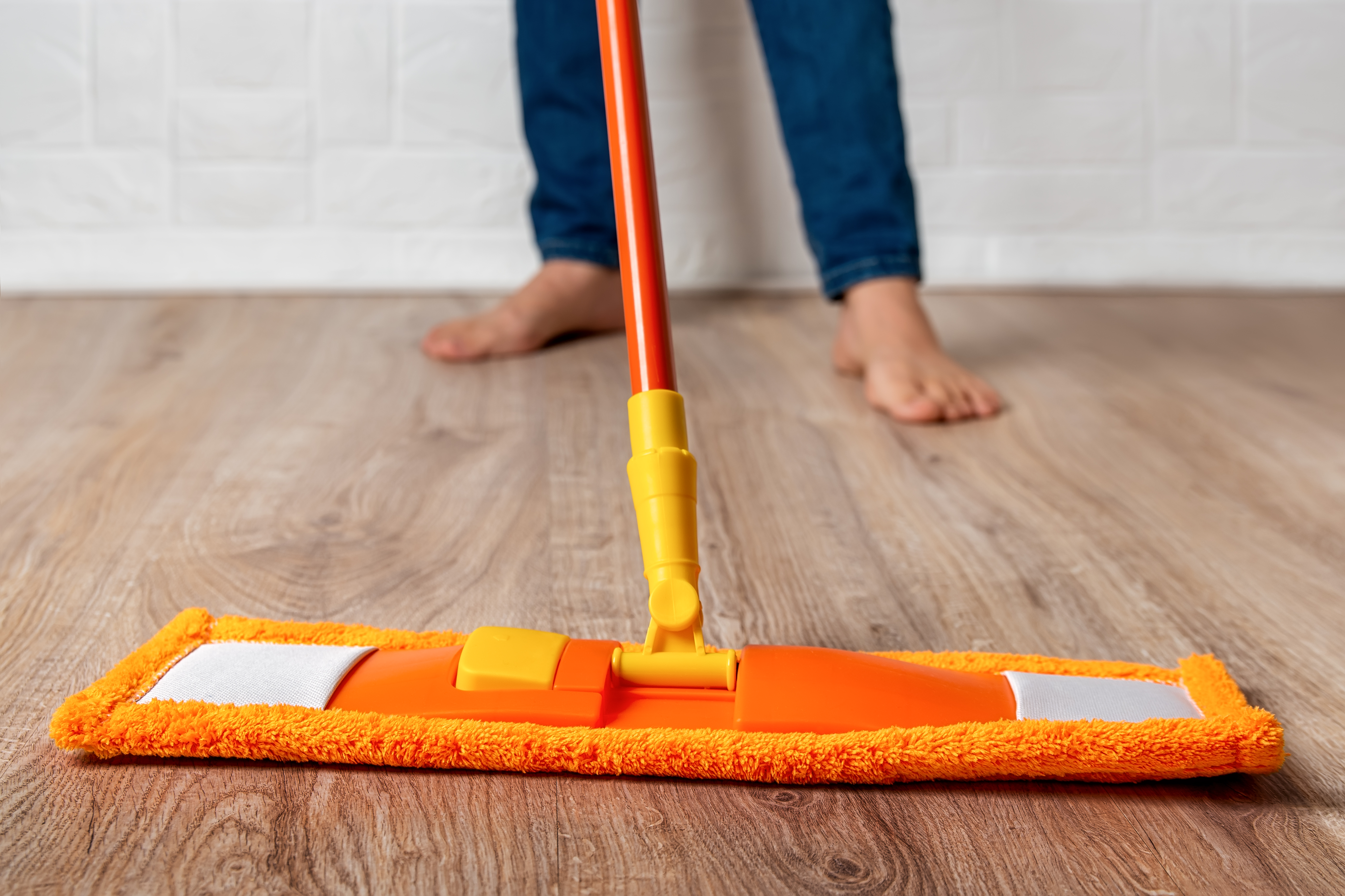 Barefoot woman mopping the floor. Female cleaning the wooden laminate floor with orange microfiber mop at home. Homework routine, housekeeping, cleaning concept. Selective focus. Barefoot woman mopping the floor. Female cleaning the wooden laminate floor with orange microfiber mop at home. Homework routine, housekeeping, cleaning concept. Selective focus.