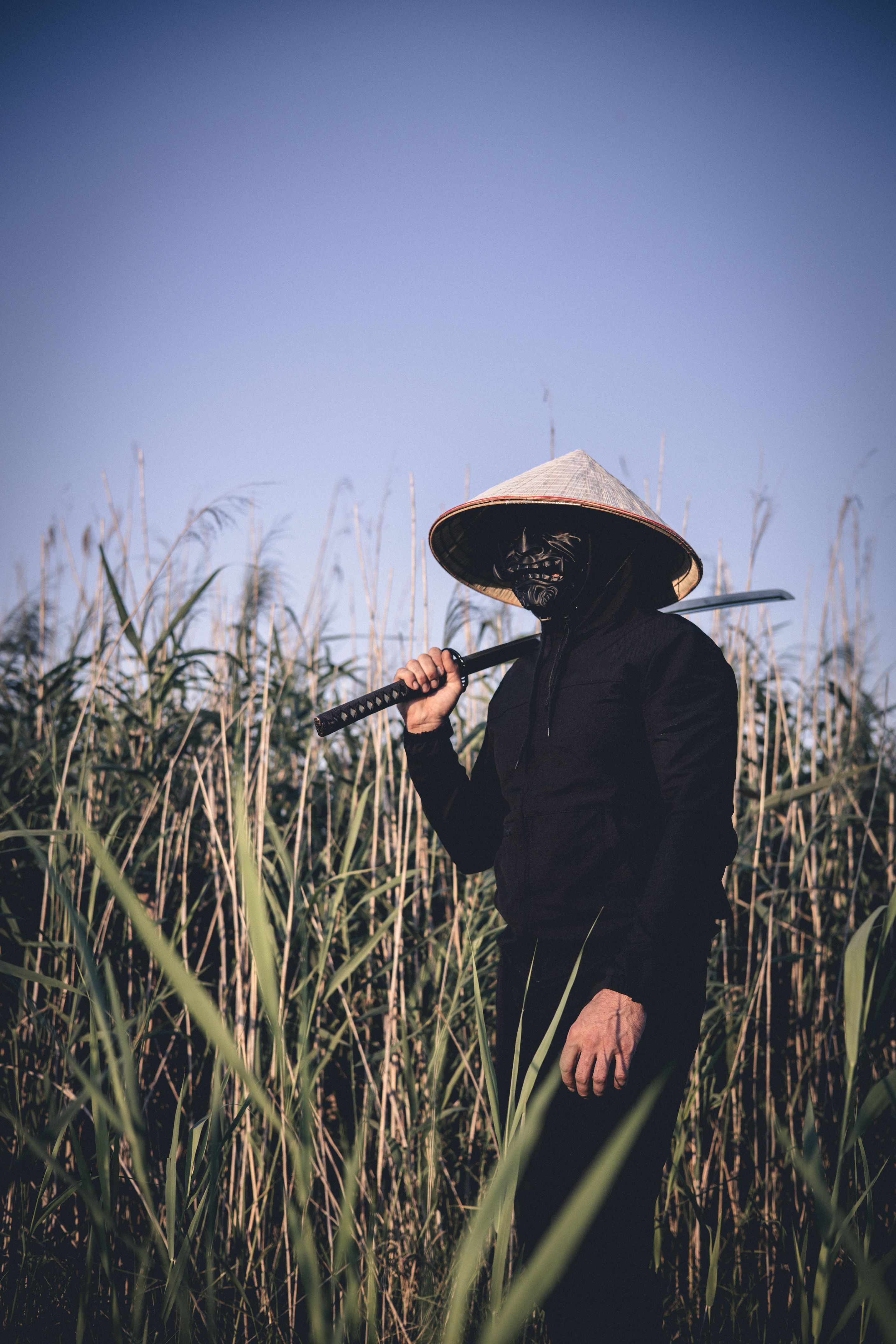 Samurai with katana against reed background. A mysterious masked samurai warrior carrying a traditional katana sword in a serene bamboo field during sunset, creating a dramatic and enigmatic scene.