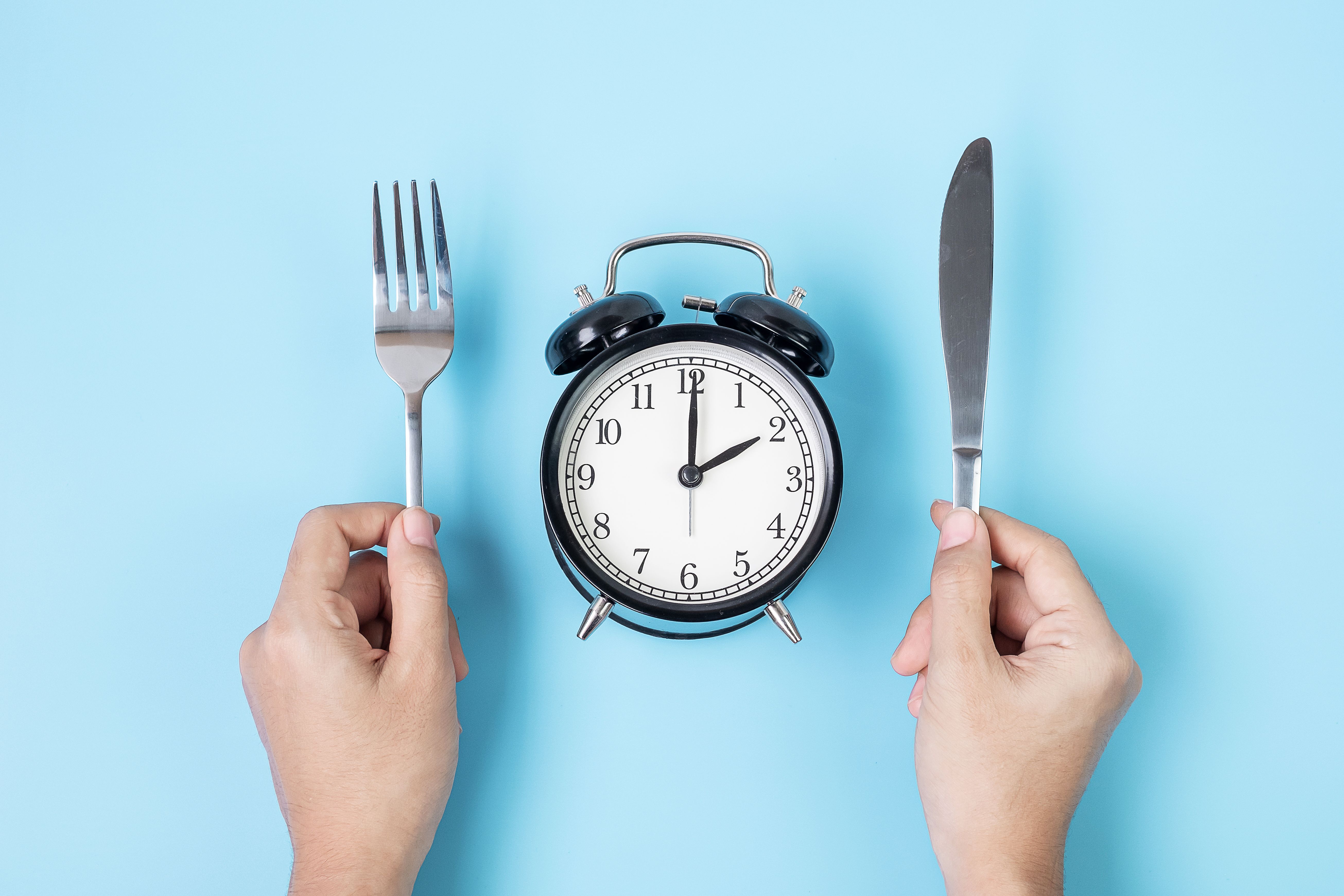 Hands holding knife and fork above alarm clock on white plate on blue background. Intermittent fasting, Ketogenic dieting, weight loss, meal plan and healthy food concept Hands holding knife and fork above alarm clock on white plate on blue background. Intermittent fasting, Ketogenic dieting, weight loss, meal plan and healthy food concept
