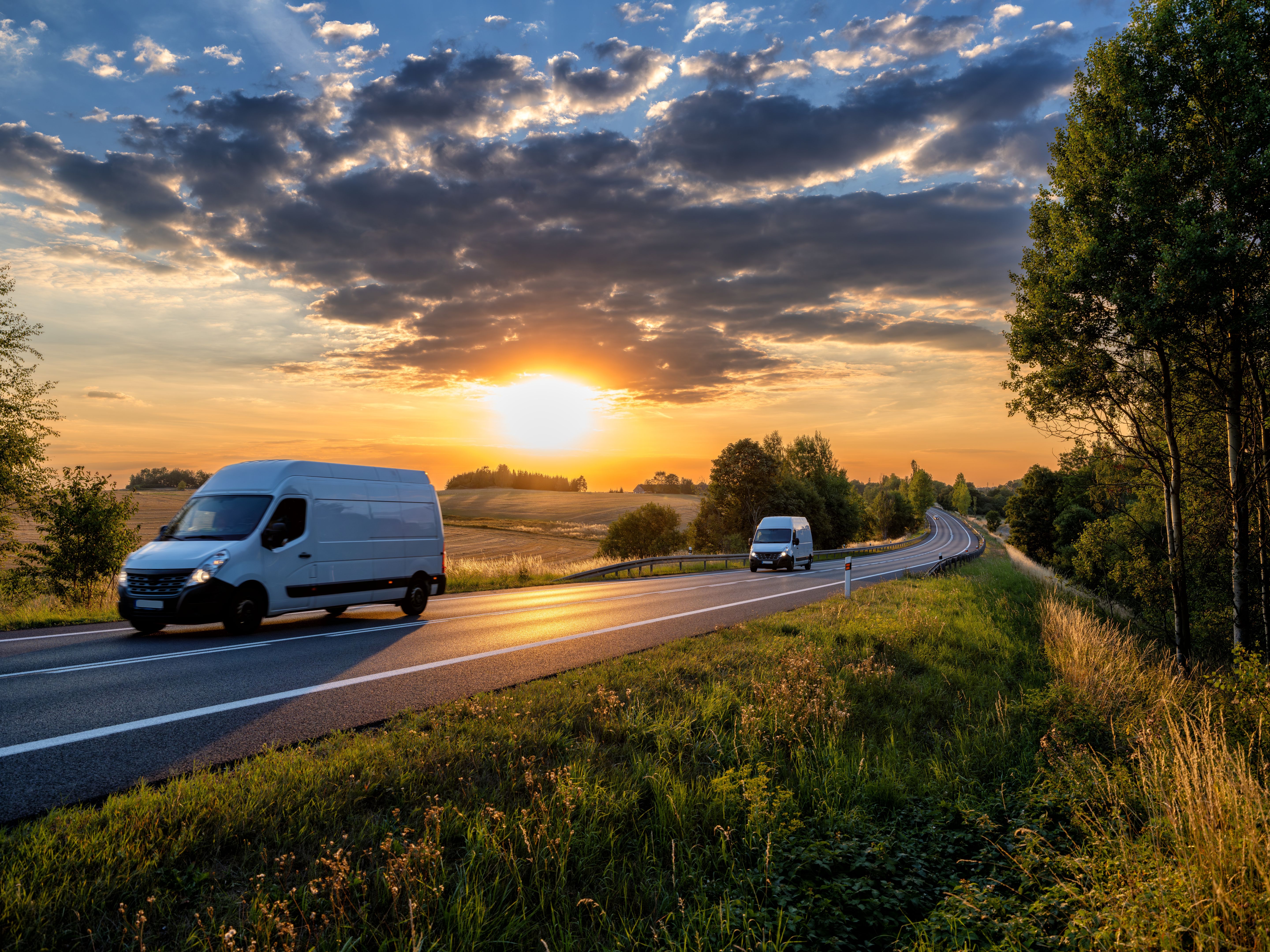 Two white fast delivery vans driving on the asphalt road in rural landscape at sunset with dark cloud