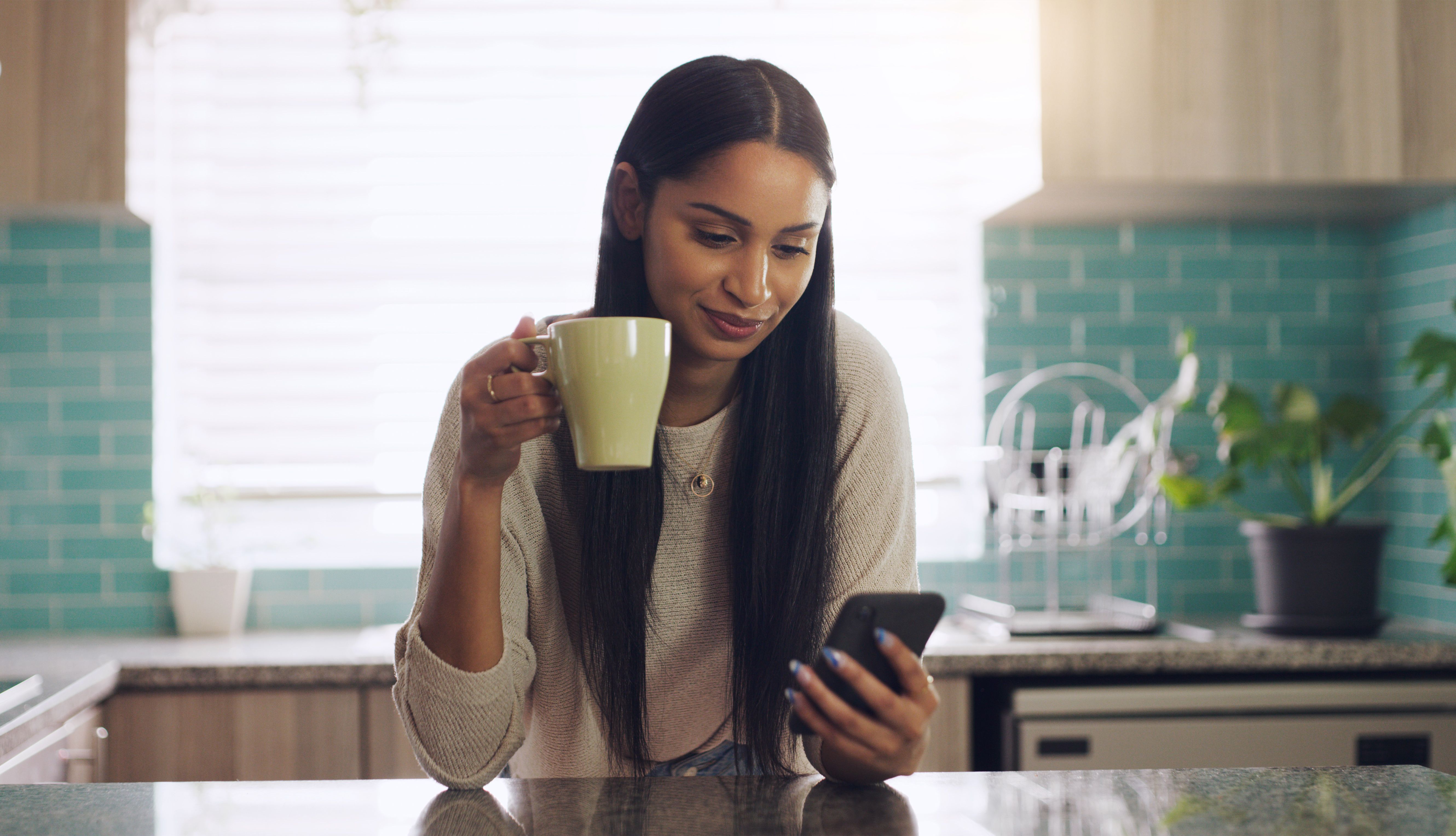 Shot of a young woman drinking coffee while using her smartphone at home Shot of a young woman drinking coffee while using her smartphone at home