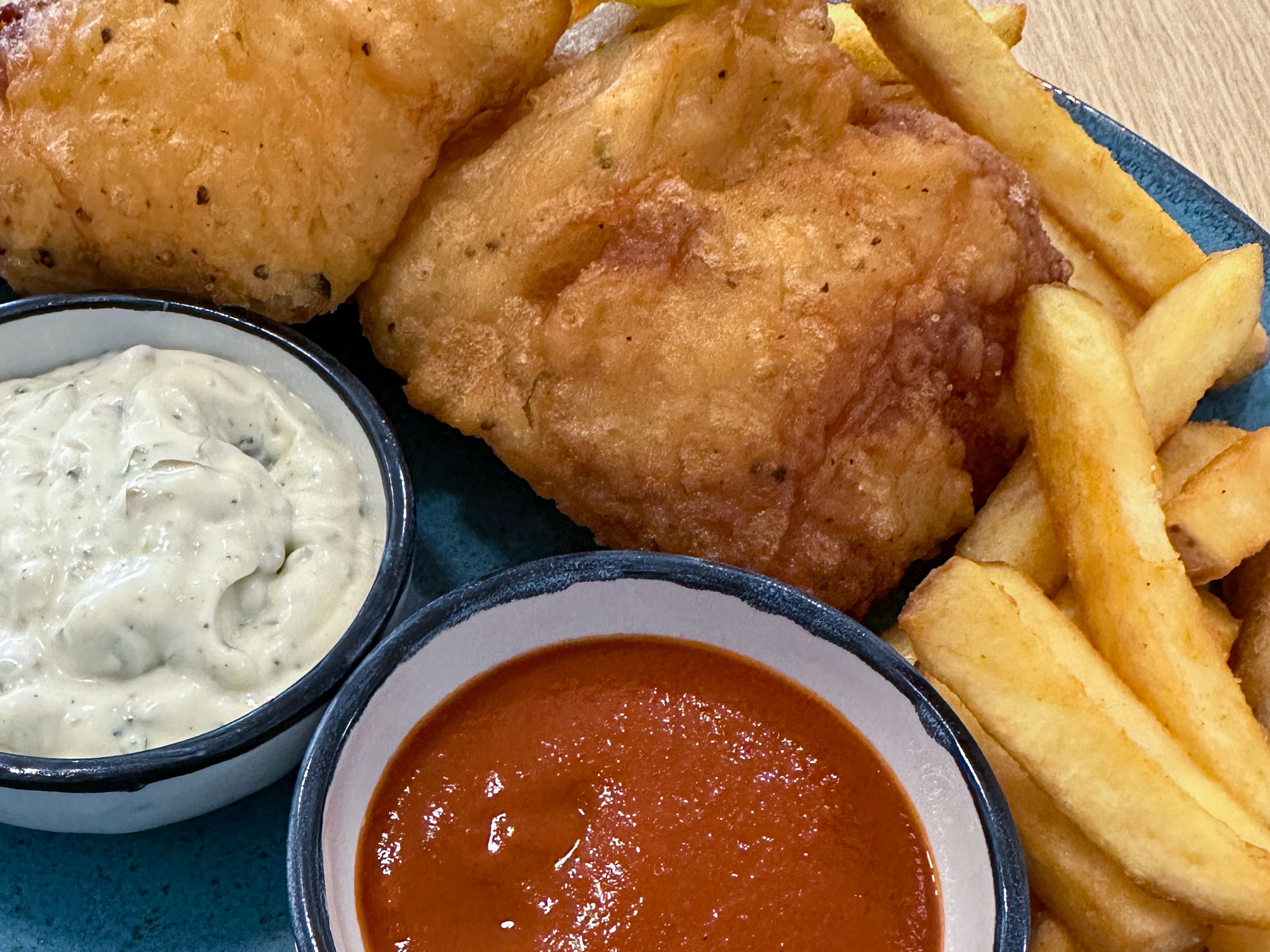 Full frame image of homemade battered cod served with French fry chips on blue plate with ramekins of tomato ketchup and tartar sauce, wood grain background, elevated view