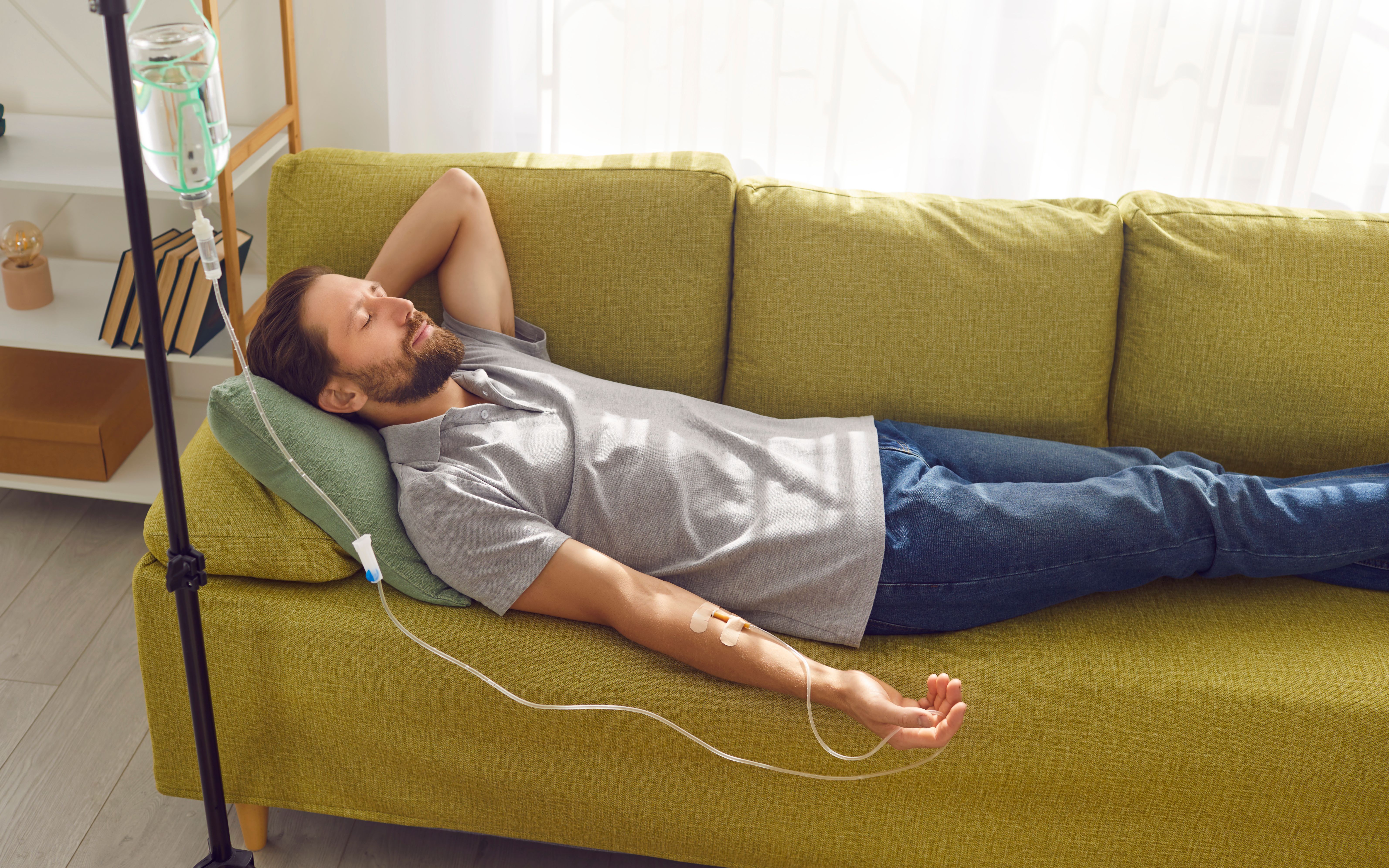 Smiling man lying on the couch at home while receiving IV drip infusion and vitamin therapy. Smiling man lying on the couch at home while receiving IV drip infusion and vitamin therapy.
