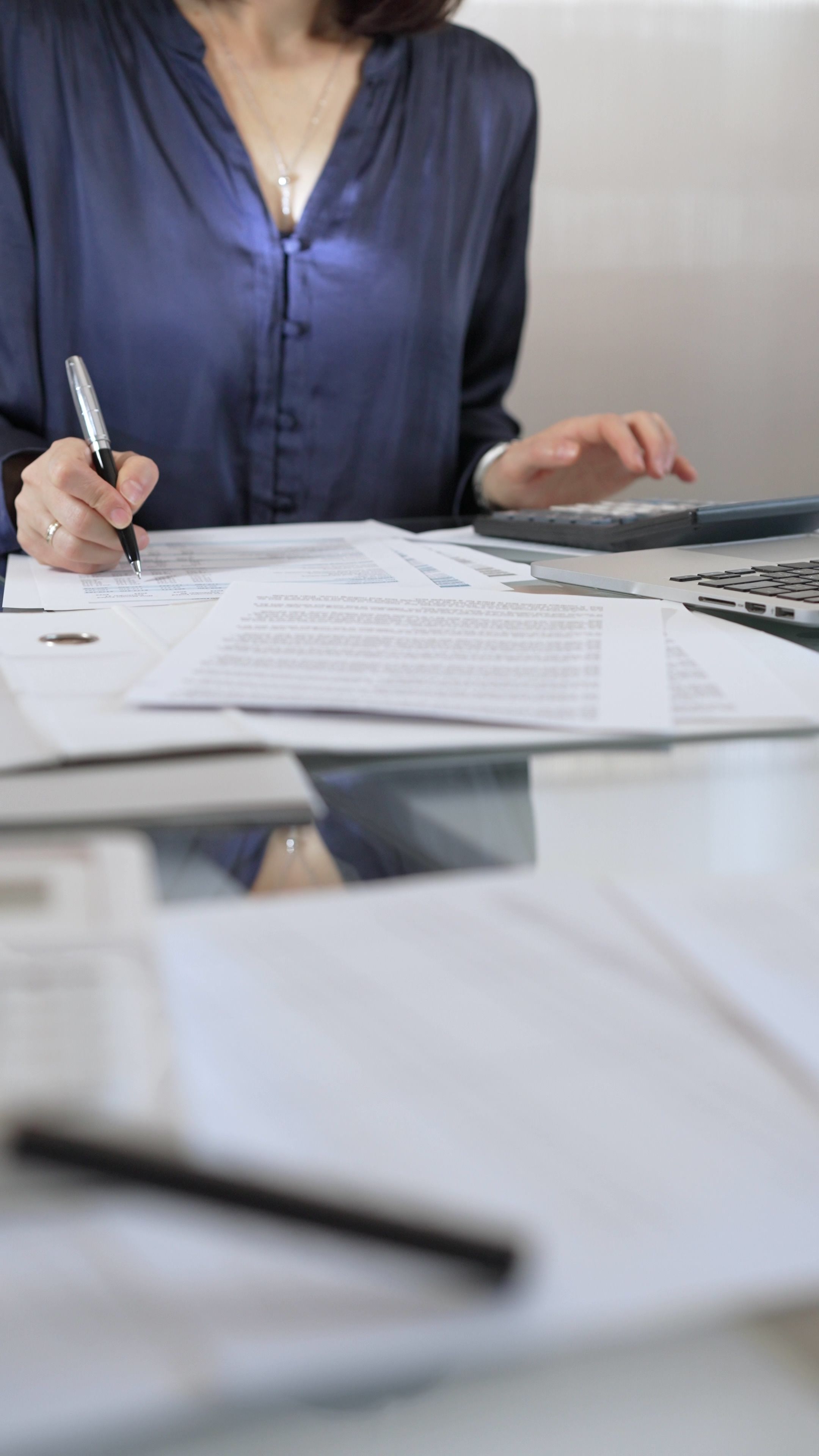 Businesswoman wearing blue dress is working with laptop, calculator and documents in a professional office setting. Low lighting, close-up vertical view. Business people, audit concept