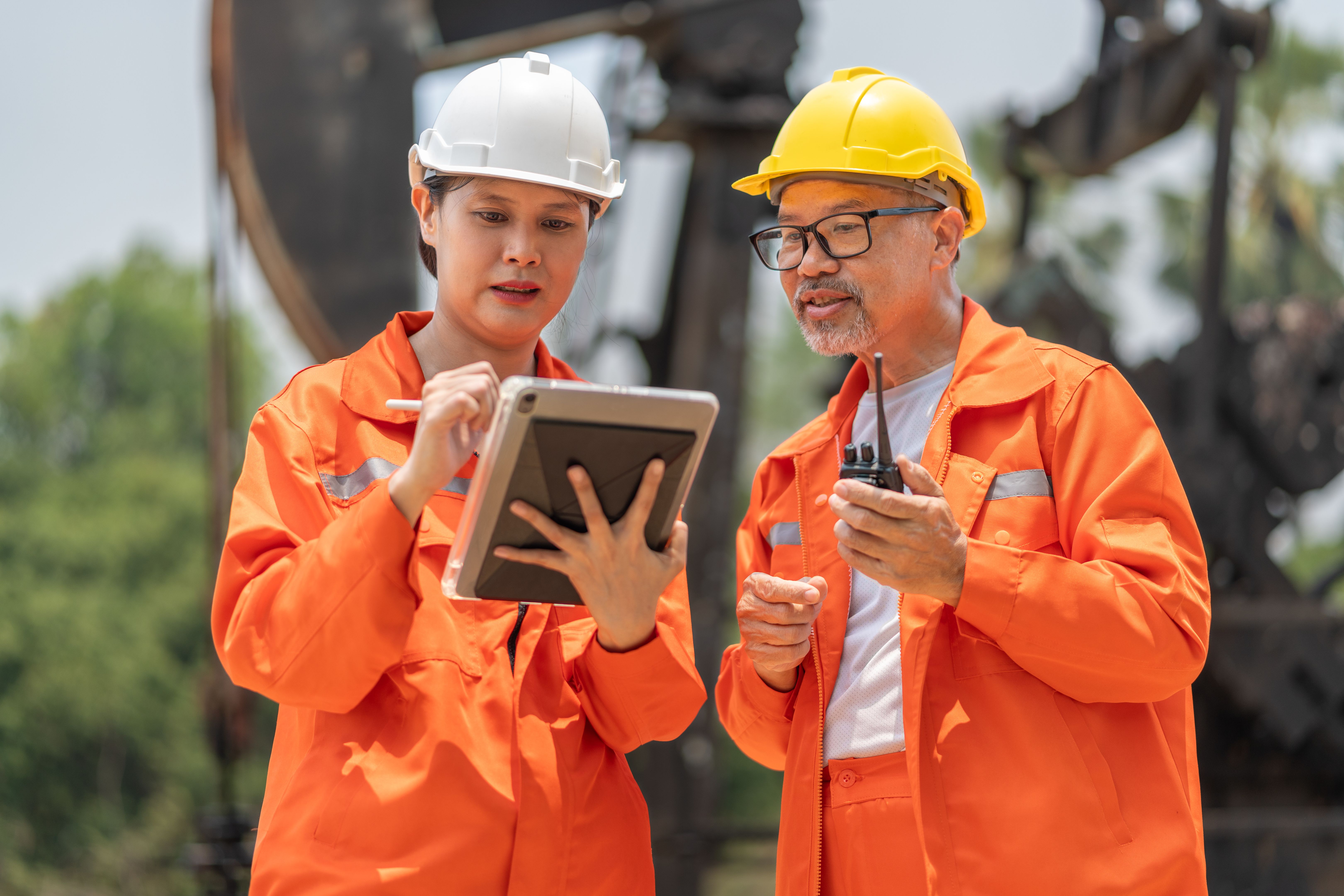 Female engineer wearing white hardhat and orange jumpsuit holding tablet working with a middle age male technician while both standing in front of an oil driller