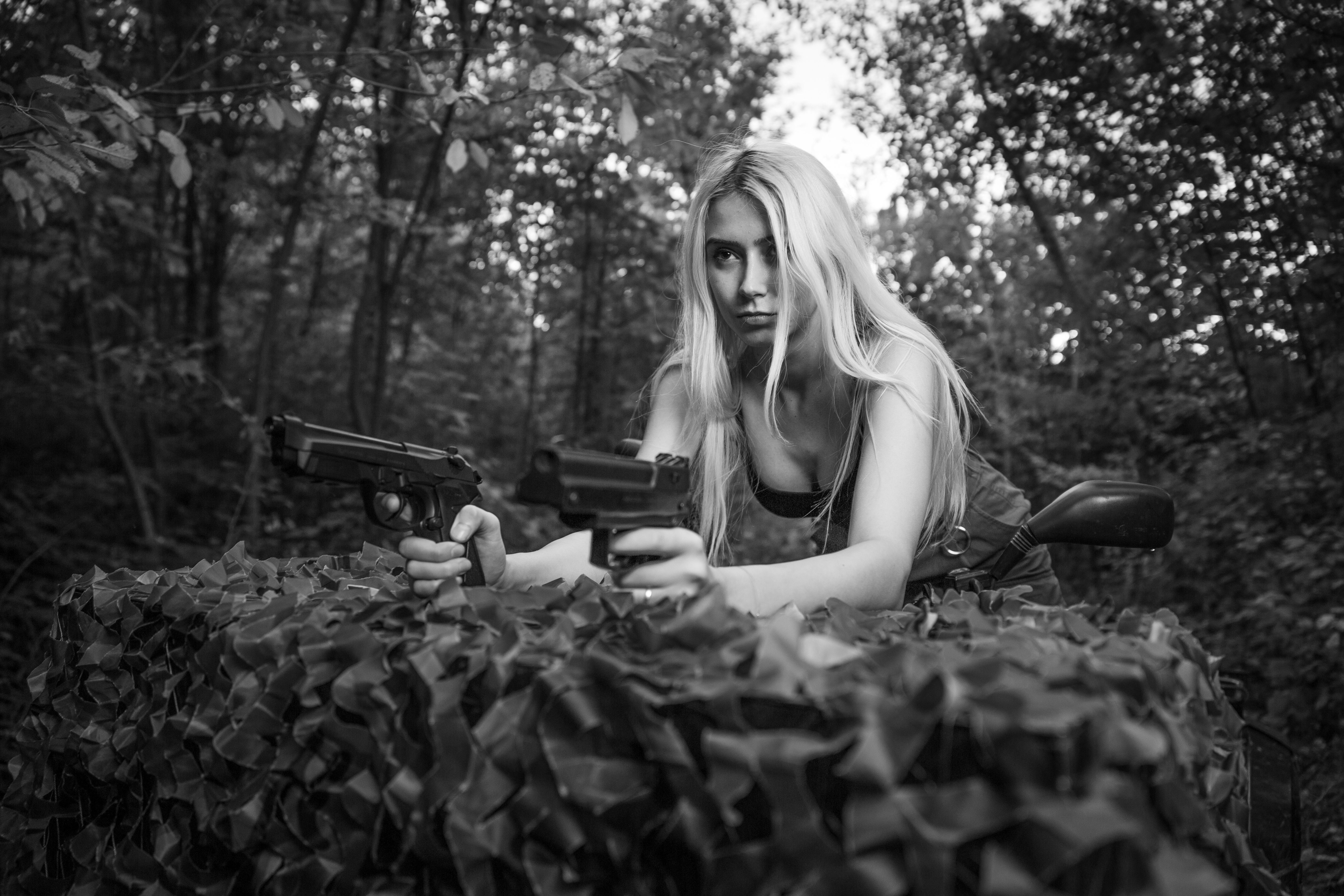 Grayscale of a Caucasian white blonde woman with guns on the quad bike in the green forest
