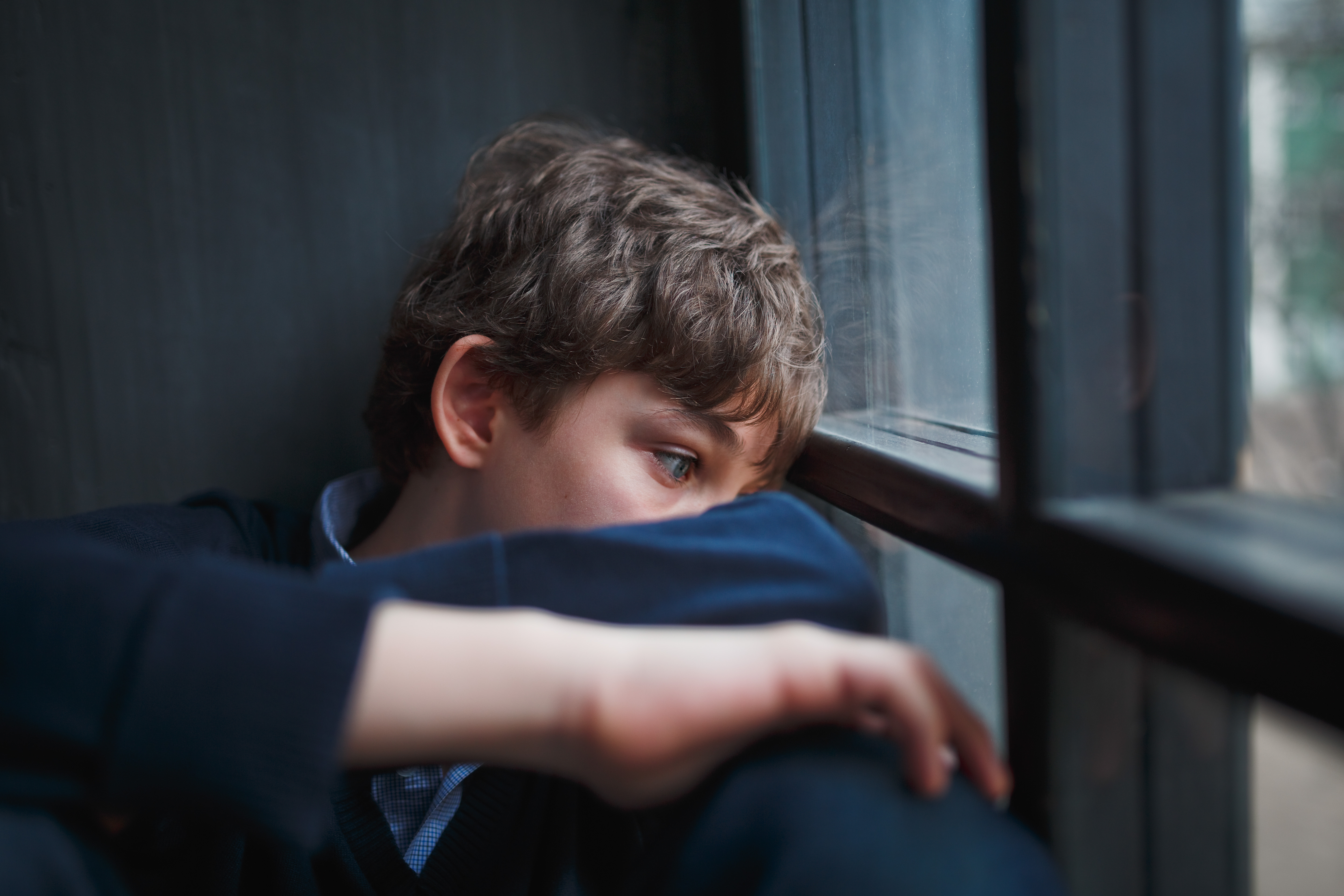 Pensive sad boy teenager in a blue shirt and jeans sitting at the window and closes his face with his hands. Pensive sad boy teenager in a blue shirt and jeans sitting at the window and closes his face with his hands.