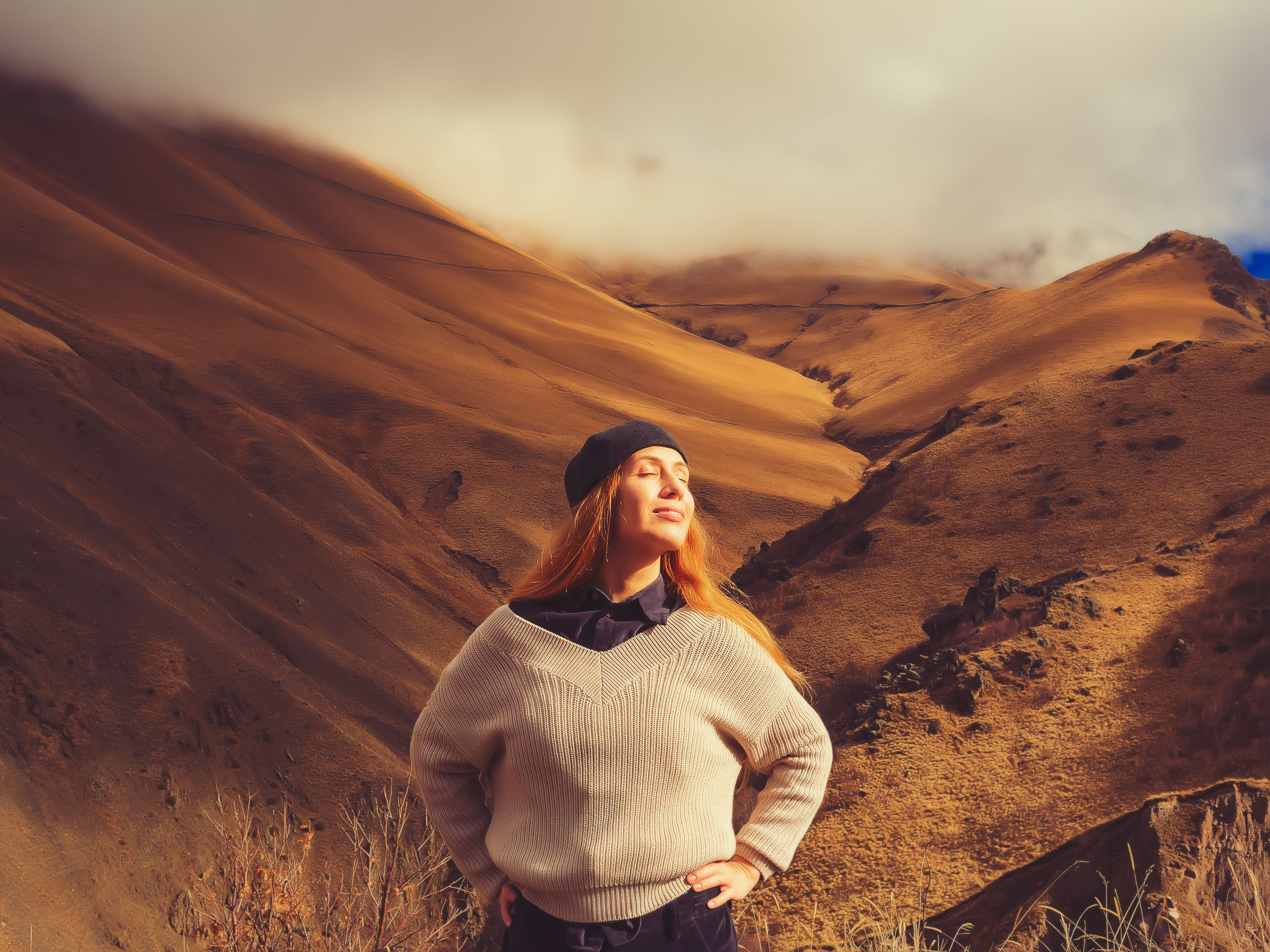 A smiling woman, closing her eyes, stands in a mountain valley with clouds covering the peaks and under the rays of the sun illuminating her face