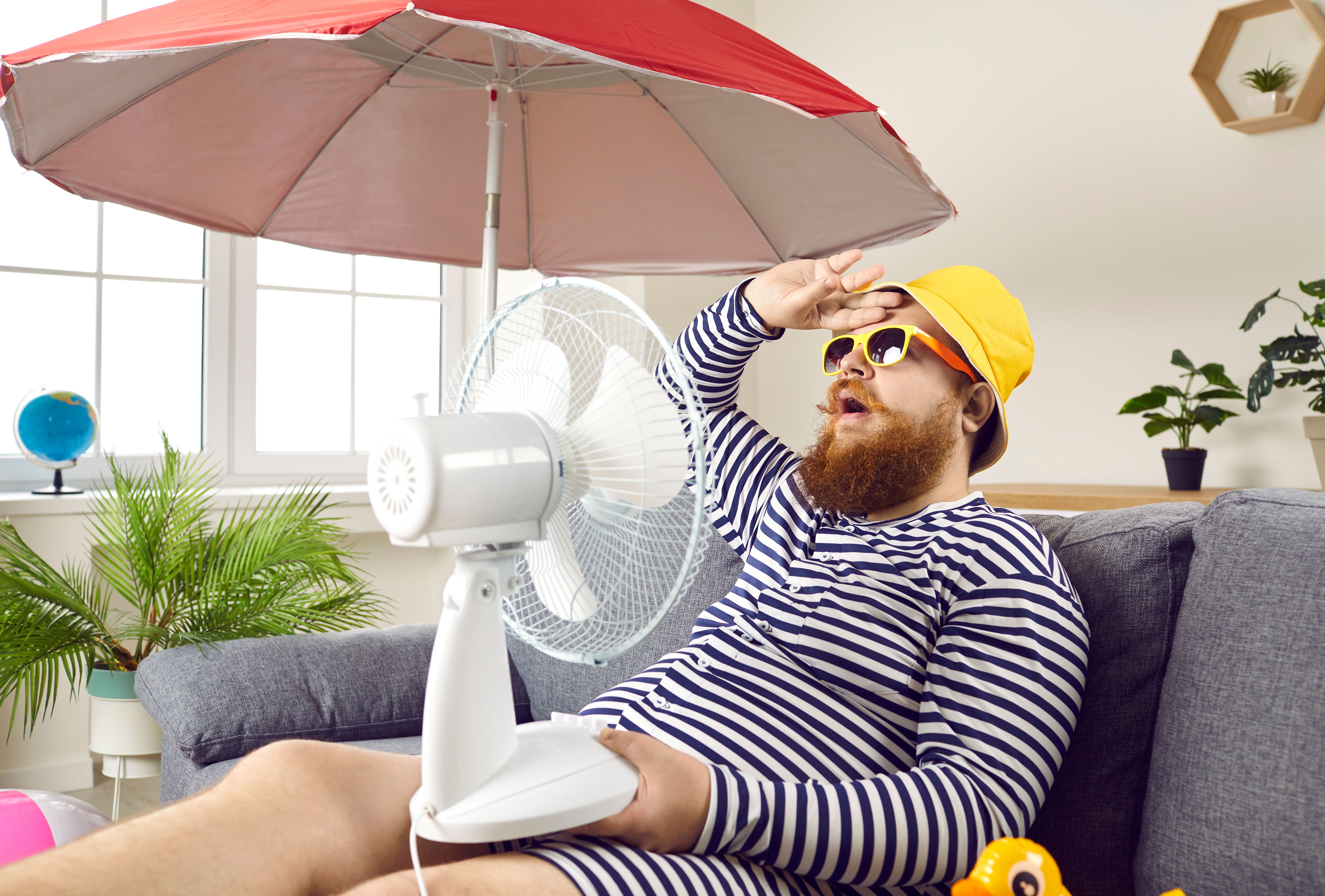 Funny man suffering from summer heat, sitting at home, sweating and holding electric fan