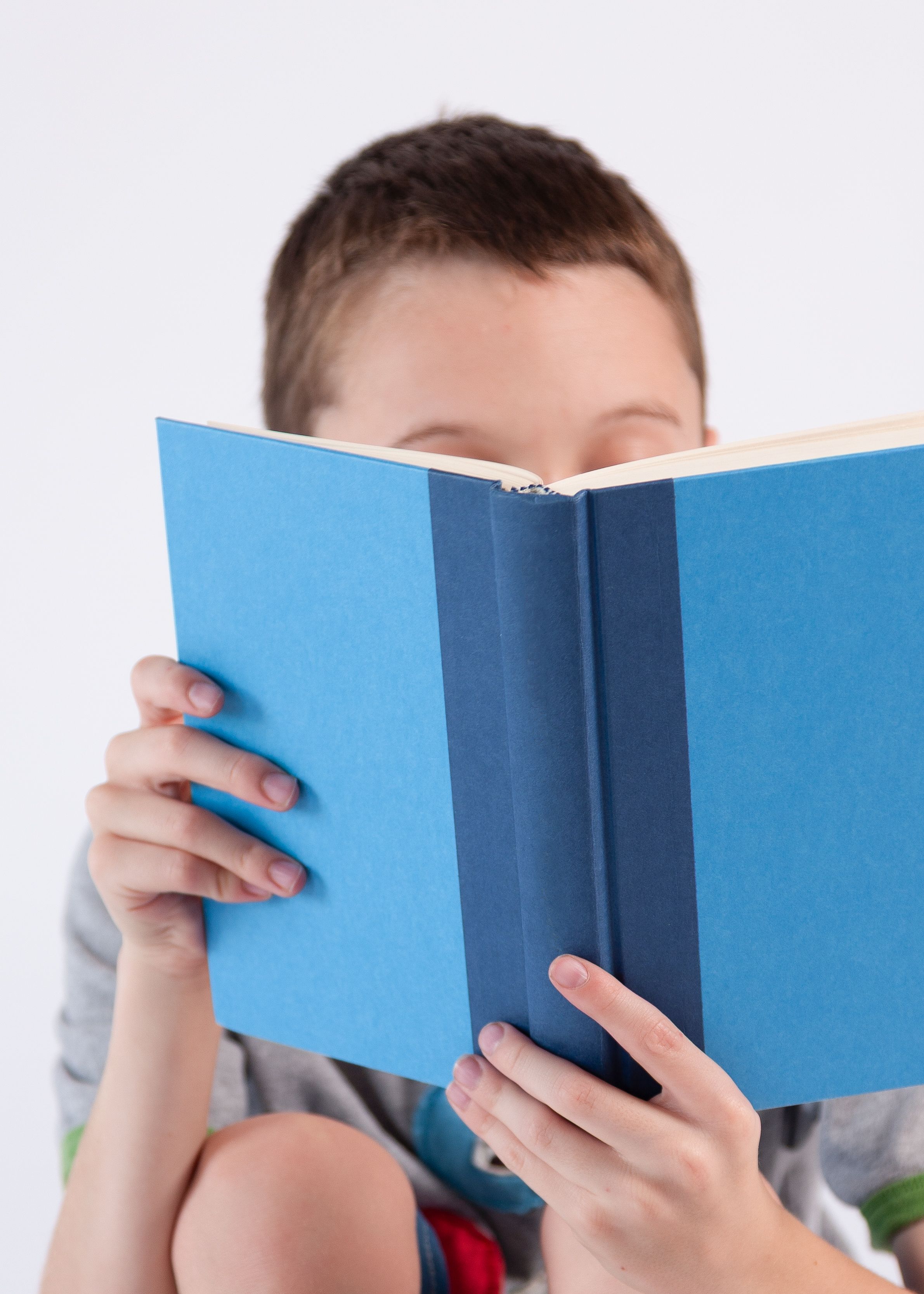 Close up of young boy reading a book with face hidden behind the book.