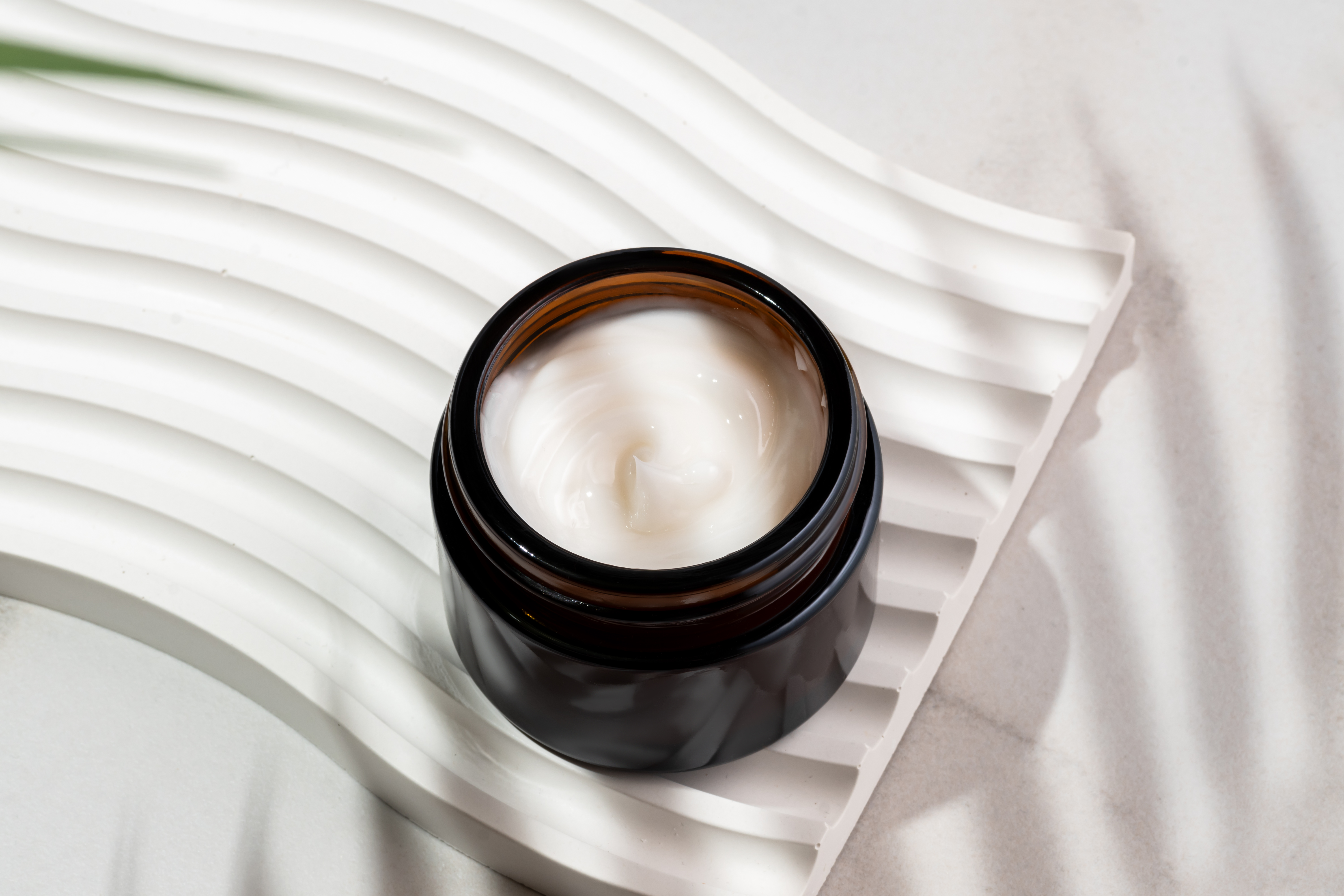 Dark glass cosmetic jar with a collagen cream on marble background with tropical leaf shadow. Natural cosmetics concept, natural essential skin care products