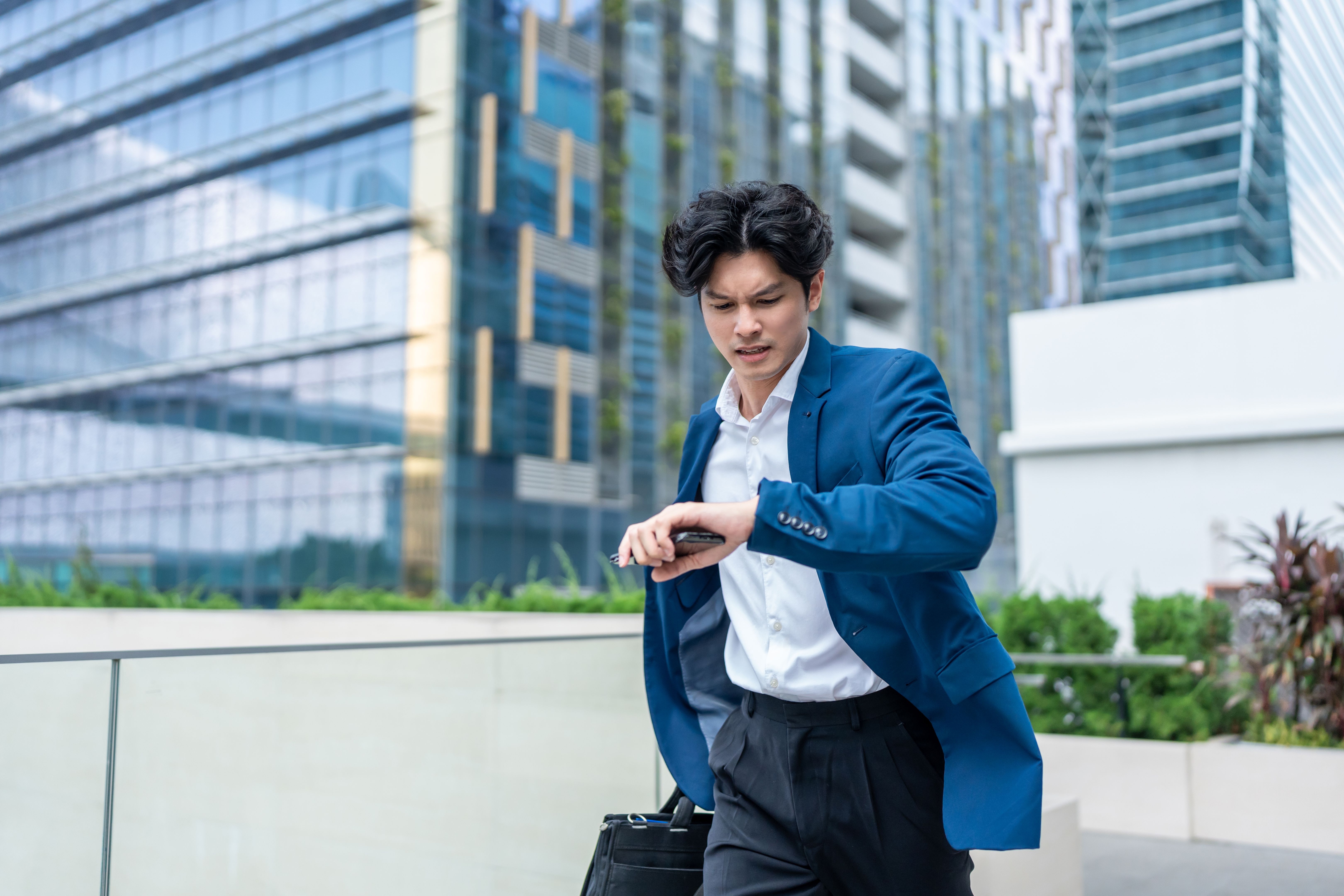 Asian young busy businessman running late to the office workplace. Attractive handsome employee man worker in formal suit holding working handbag in hurry walking outdoors in the modern urban city. Asian young busy businessman running late to the office workplace. Attractive handsome employee man worker in formal suit holding working handbag in hurry walking outdoors in the modern urban city.