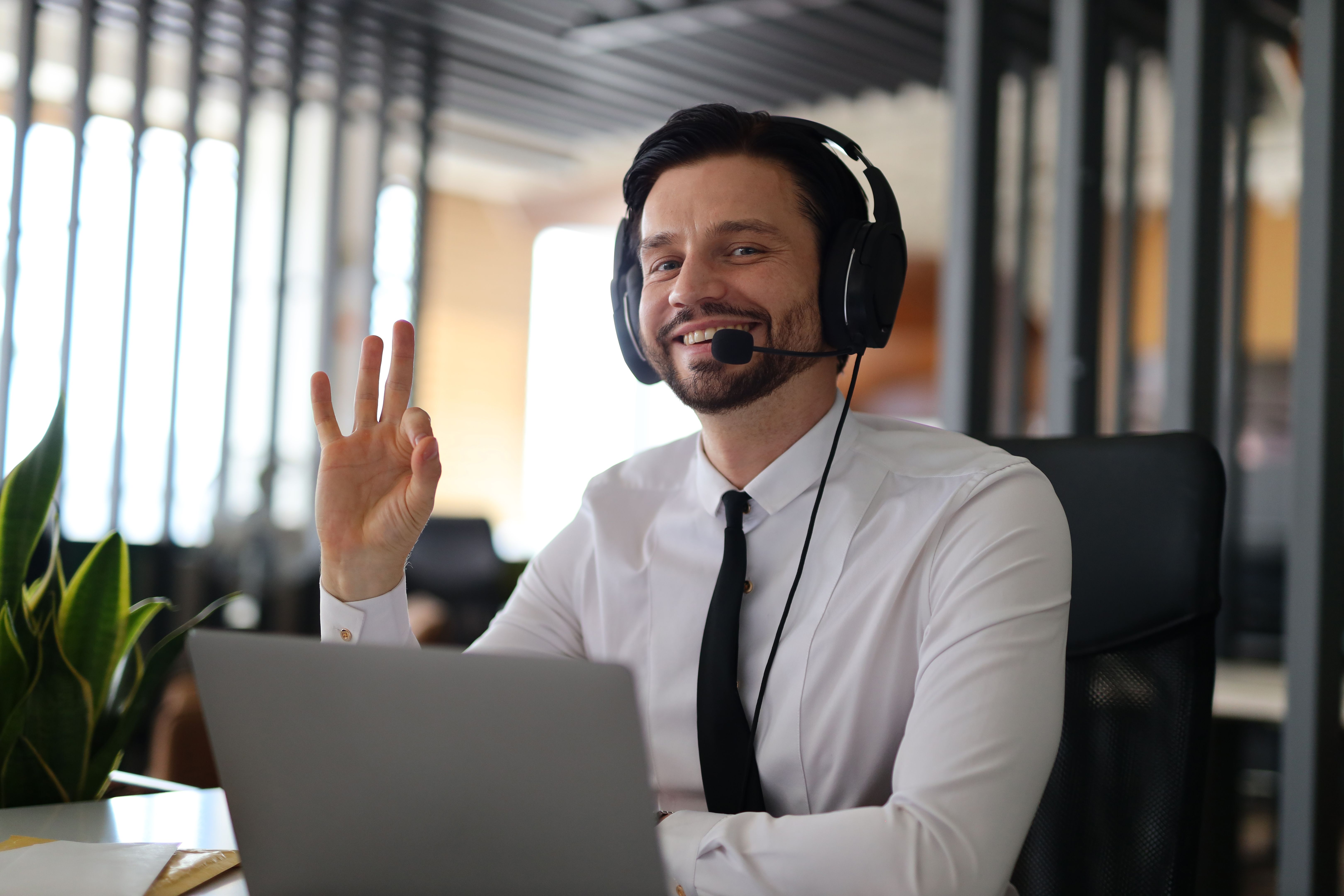Ein Mann mit Headset und Krawatte lächelt, während er mit einem Laptop spricht. Er befindet sich in einem professionellen Umfeld und hat Spaß an seiner Arbeit