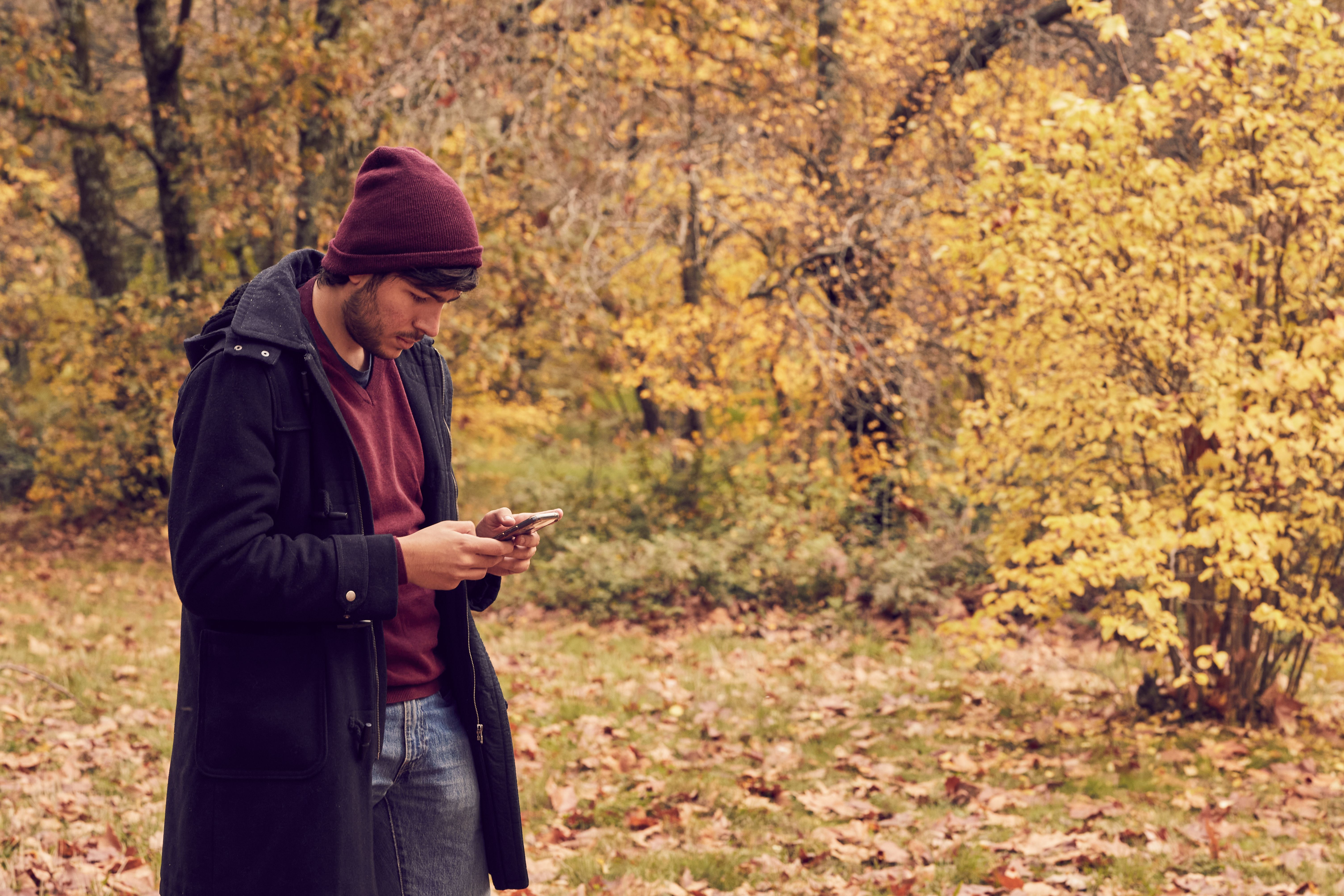 White caucasian man in the forest dressed a long coat and cap looking at smartphone. Autumn colors