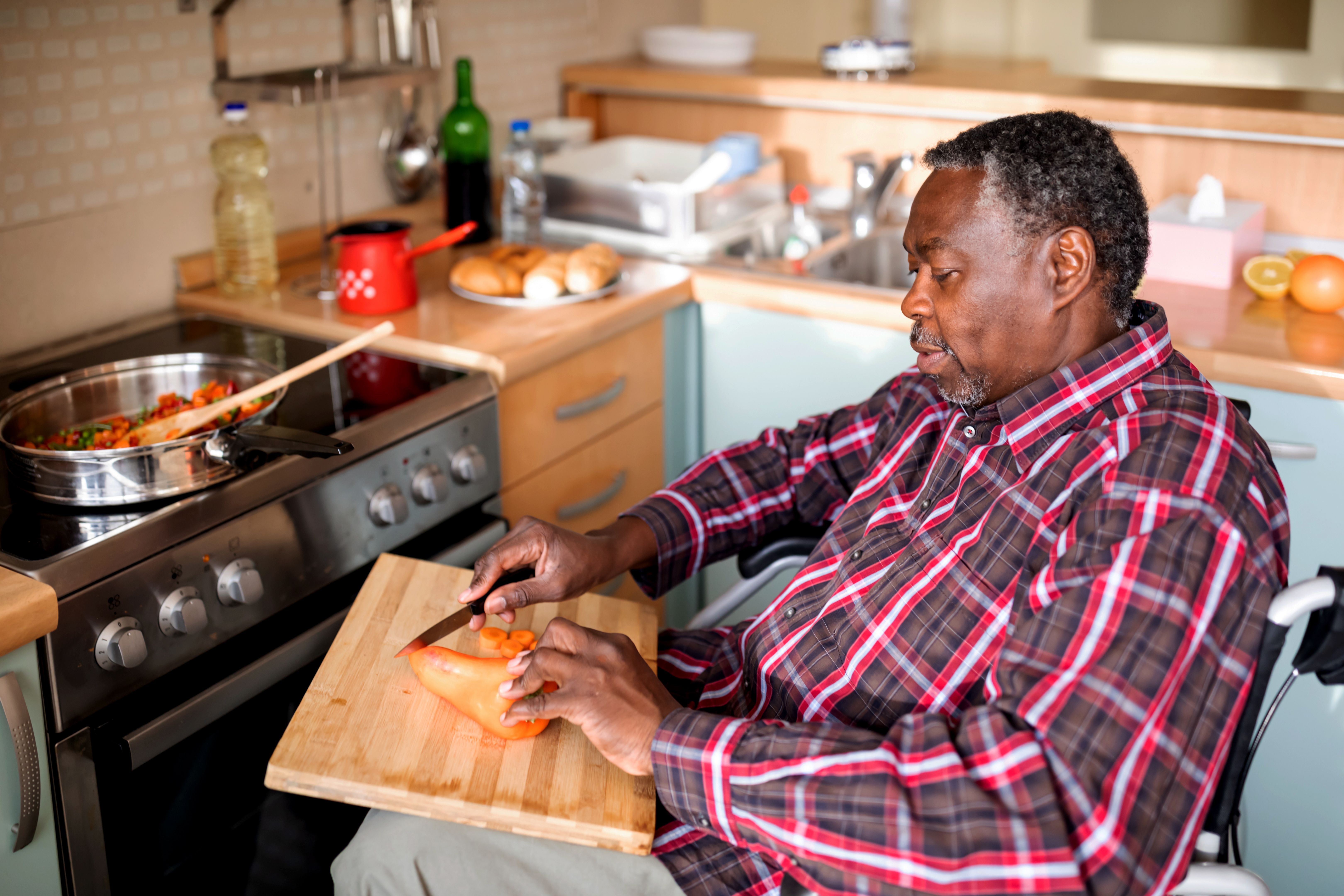 Senior Man with mobility handicap slicing red pepper using sharp knife in kitchen. Disabled paralyzed handicapped man with walking disability integrating after an accident.